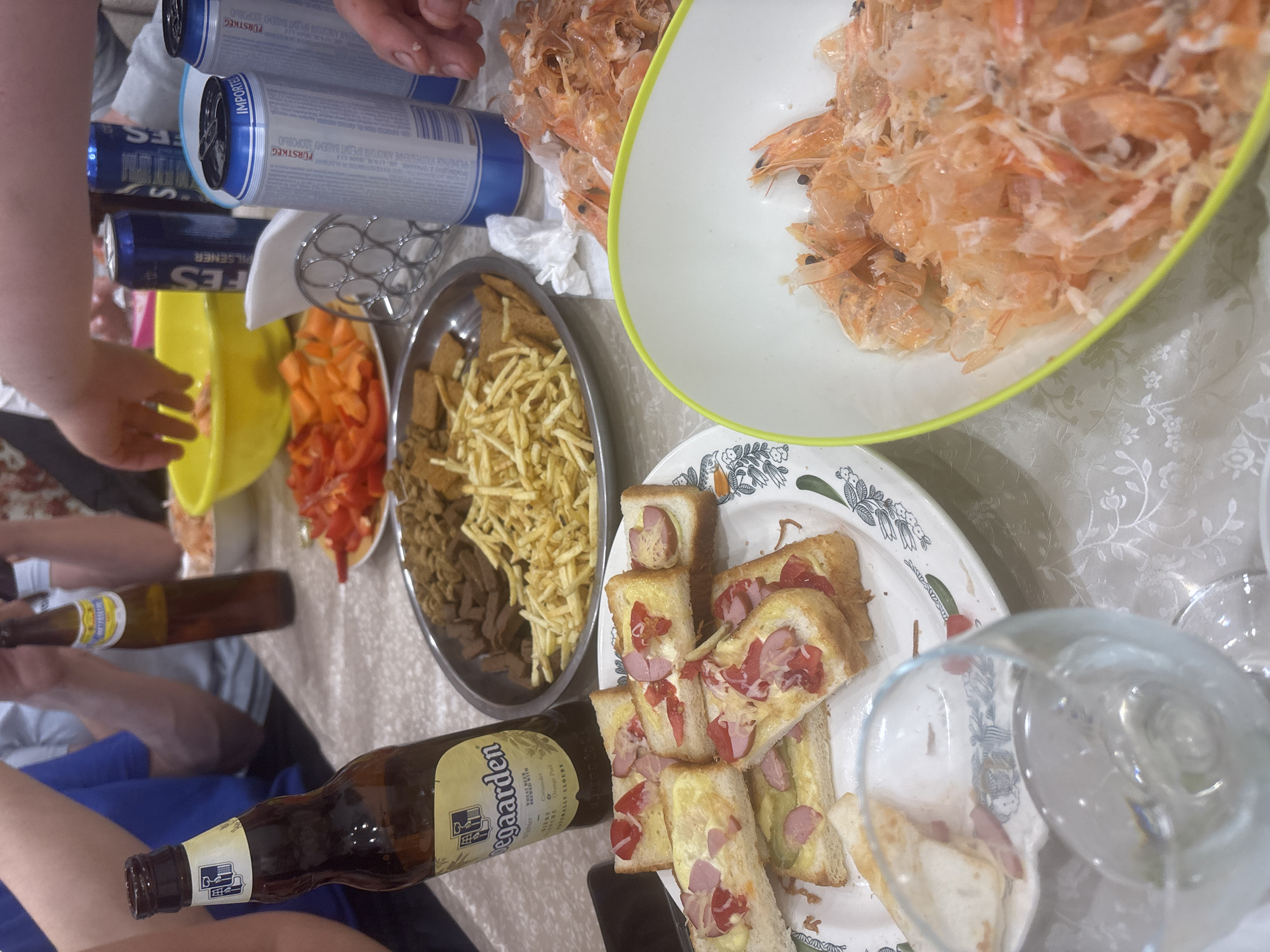 The image shows a table laden with various foods and drinks. There are several containers of what appear to be shrimp shells, along with bowls of snacks such as chips and sliced vegetables. Additionally, there's a plate of sandwiches, some beer bottles (including one labeled "Hoegaarden"), and cans of "FES" beer. A few people are visible around the table, seemingly in the act of eating and drinking.
The OCR tags suggest the location is possibly in Russia, given the Cyrillic text, and the presence of "FES" beer may indicate a connection to Ferma, as "FES" is mentioned in the OCR and associated with Russia. However, there's no definitive visual evidence in the image to confirm the precise location is the city of Ferma.