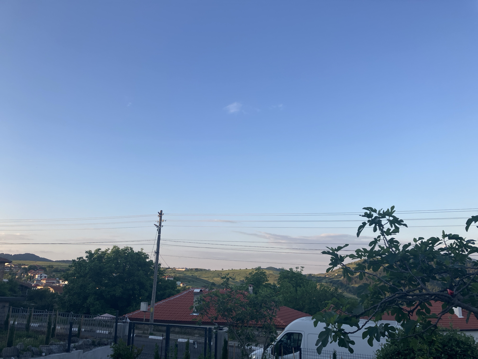 The image depicts a daytime view in Komuniga, Bulgaria. In the foreground, there are rooftops of buildings with red tiles, trees, and a white van. Power lines stretch across the midground, with rolling hills and scattered vegetation visible in the distance. The sky is blue with a small patch of clouds. A utility pole is seen amidst the buildings and trees.
