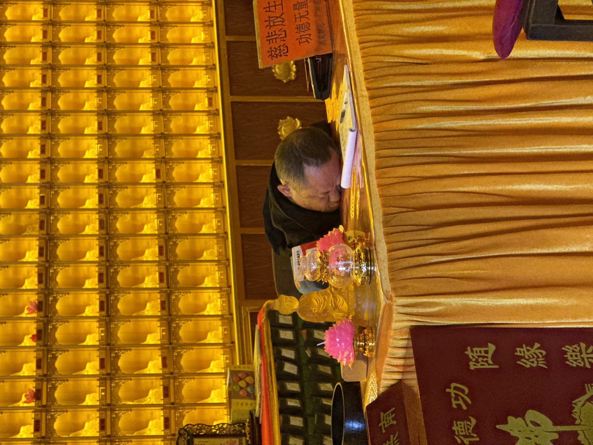 A person with short, graying hair bows their head to a table draped in gold cloth in what appears to be a Buddhist temple or shrine. The table is adorned with pink flowers, a small golden Buddha statue, glass containers, and a red book with Chinese characters. A notebook sits open on the table. Behind the person is a grid-like wall filled with small compartments illuminated from within, possibly containing individual offerings or memorials. A sign with Chinese characters is visible above the person's head. The scene appears to be indoors and has an overall golden hue due to the lighting and decor. The location is likely Yongding District, China.