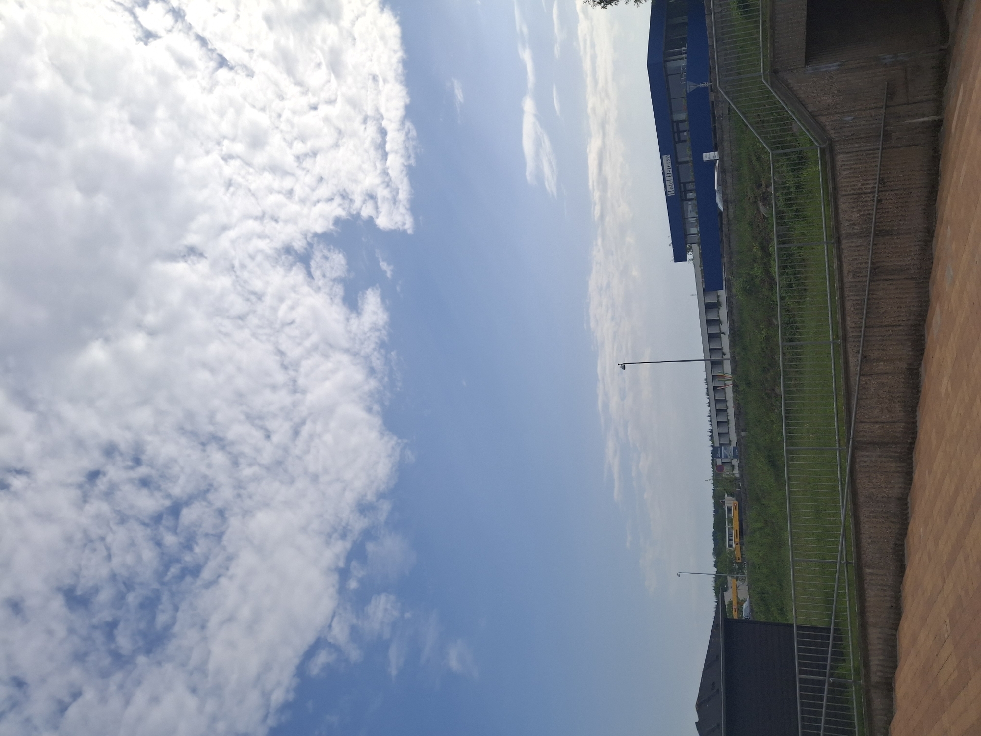 The image shows a view from a slightly elevated position in Aabenraa, Denmark. The sky occupies the left side of the image, displaying a mix of blue sky and white, puffy clouds. On the right side, a brick structure with a metal railing is visible, suggesting a pathway or stairway. Behind the railing, there's a grassy slope leading down to what appears to be an urban landscape with buildings, including the "Hotel Ostsee". A street with vehicles, including a bus, can be seen in the background. The scene is captured in daylight.
