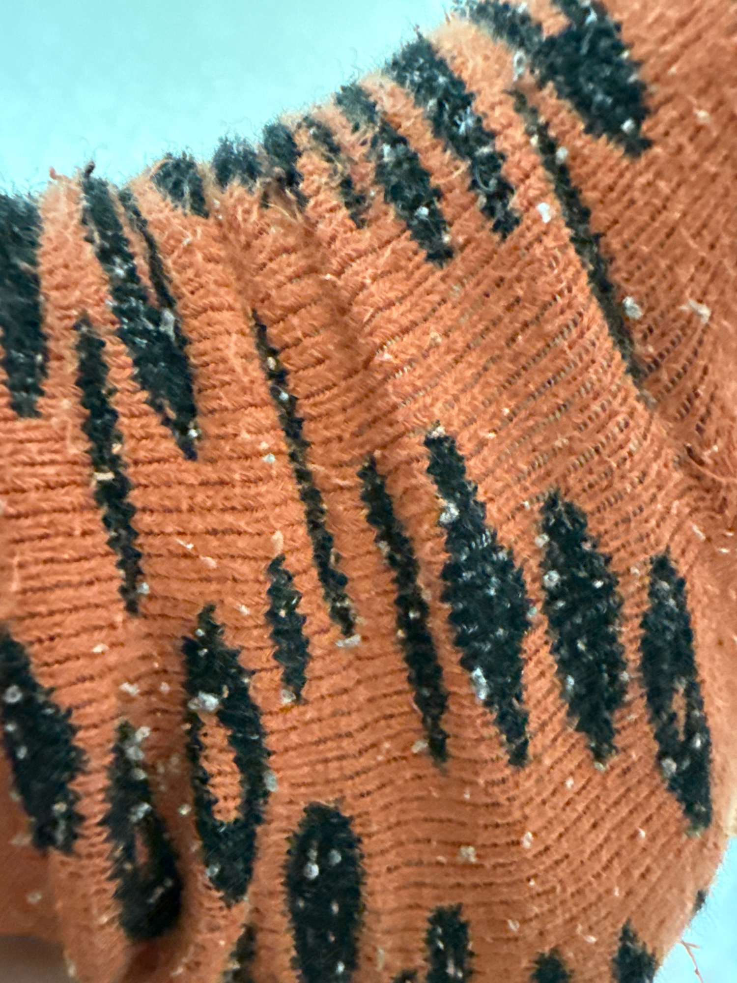 A close-up, overhead shot focuses on a textured, orange fabric with black tiger-like stripes. The fabric appears to be a sleeve or similar garment, with visible stitching and a slightly fuzzy surface. Numerous small, white specks are scattered across the orange sections of the fabric, appearing to be lint or debris. The background is a bright, solid blue. The scene is devoid of any human presence or discernible location within Zavolzhye, Russia.