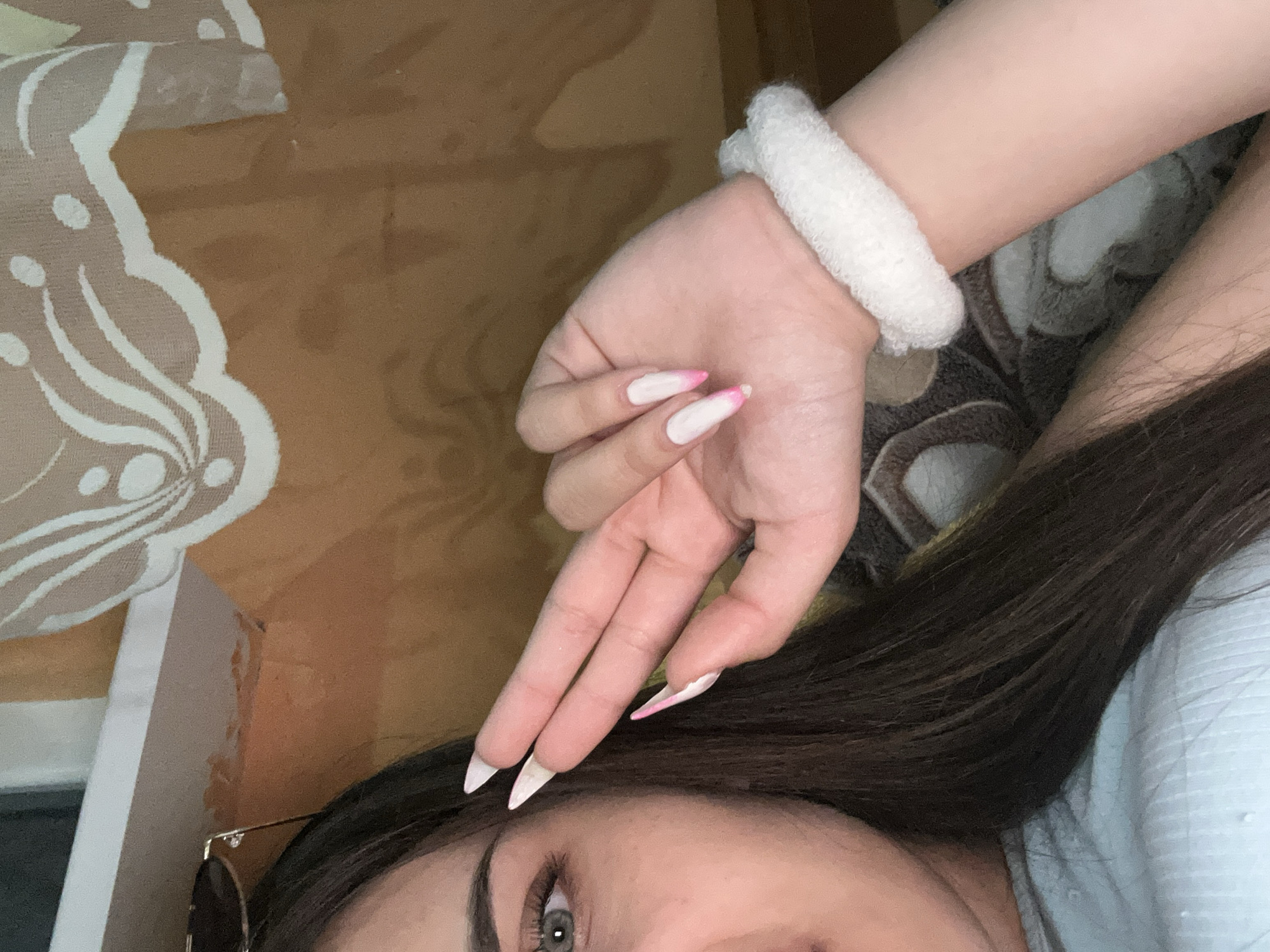 A close-up, partial view shows a person's face, hand, and arm. The person has long, dark brown hair and striking green eyes. Their hand is positioned in front of their face, with fingers extended and adorned with long, pointed white nails with pink tips. A fluffy white scrunchie is visible around their wrist. The background consists of a textured, brownish-orange fabric with a white floral pattern overlaid on a section. The lighting suggests an indoor setting, likely a domestic environment. There is no discernible information in the image that would allow for the determination of its location within the city of Shumen, Bulgaria.