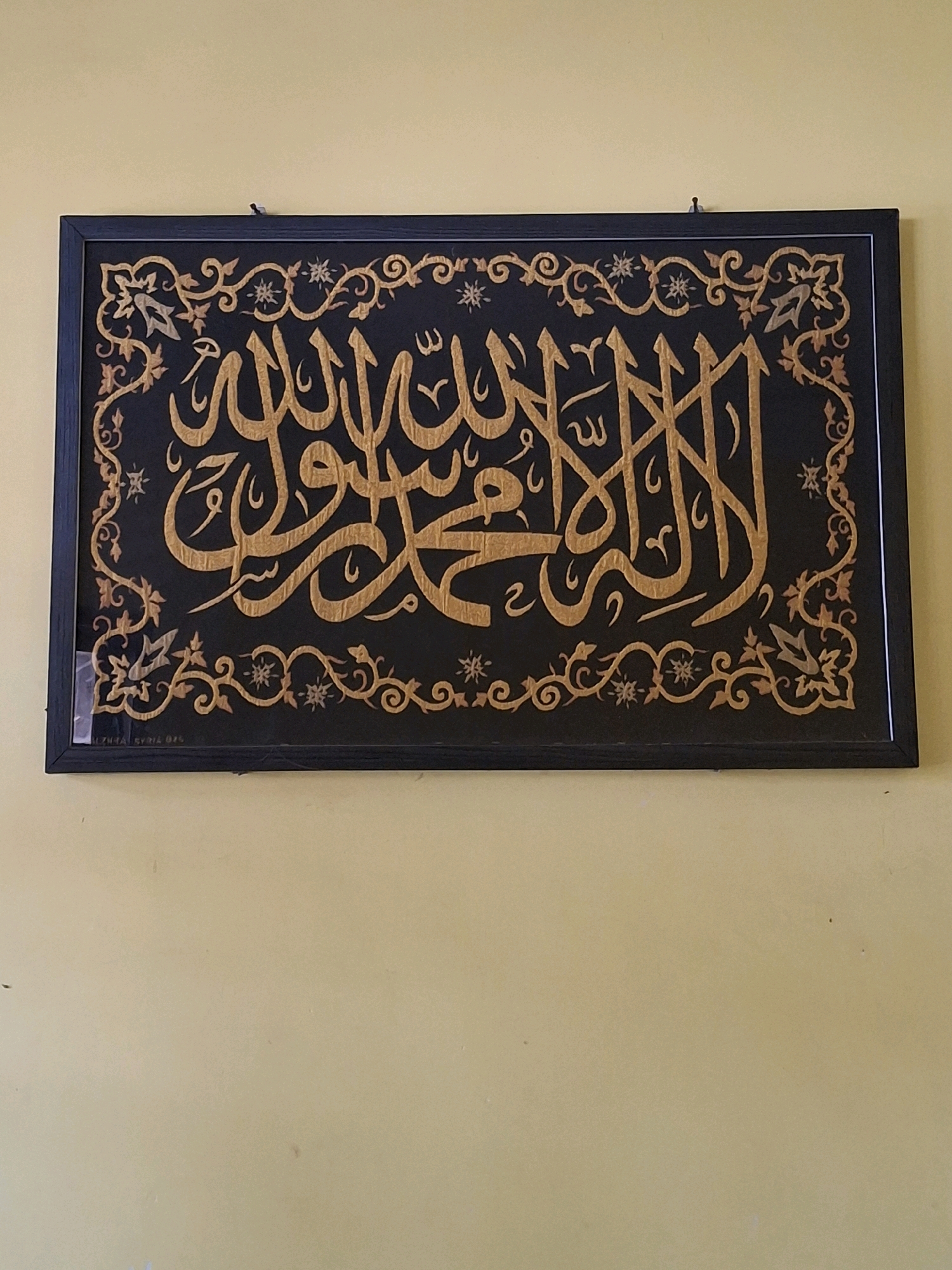 The image shows a framed piece of Islamic calligraphy hanging on a pale yellow wall. The calligraphy is rendered in gold on a dark background and features intricate patterns around the edges. A small nail is visible at the top center of the frame, securing it to the wall. The lower-left corner of the frame has a small white sticker or label. The wall appears to be painted and has some minor imperfections or marks. There is no indication that this scene is located in Lumajang, Indonesia.