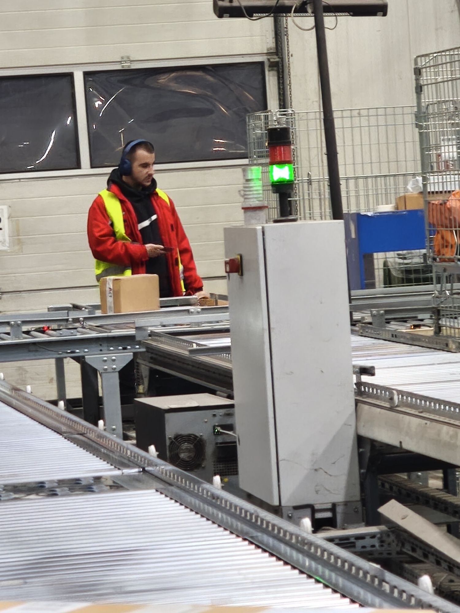 The image depicts an indoor industrial environment. A man wearing blue headphones, a black hoodie, and a red and yellow reflective jacket stands near a conveyor belt system. He appears to be holding a small black object, possibly a phone or scanner, in his hand. A cardboard box sits on the conveyor belt near him. In the foreground, a complex network of metal conveyor rollers stretches into the distance. A gray control box with a red emergency stop button and a green signal light above it is positioned near the conveyor system. Behind the man, there's a large gray door with rectangular windows covered by clear plastic. Metal cages are visible in the background. There is no definitive evidence that this scene is in Burgas, Bulgaria.