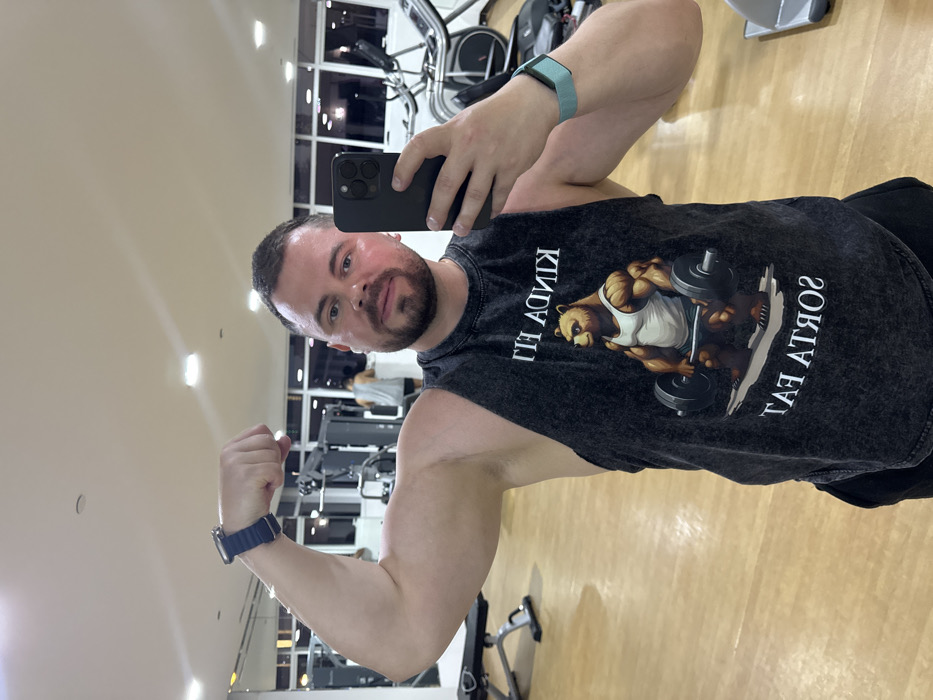 A male subject is depicted in a mirror selfie taken inside a gym, located in the city of Dubai, United Arab Emirates. The subject, a man with short dark hair and a beard, is positioned centrally, facing the camera. He is flexing his left bicep, with a dark blue smartwatch on his left wrist. He holds a dark-colored smartphone in his right hand to capture the image, displaying a light blue fitness tracker on his right wrist. He wears a dark grey, sleeveless tank top featuring an inverted graphic of a muscular bear lifting a barbell, accompanied by the text "Kinda Fit Sorta Fat." The gym environment includes light-colored flooring, recessed ceiling lights, and large windows revealing a dark exterior, indicating nighttime. Various gym equipment, including weight benches, strength training machines, and cardio equipment (an elliptical and a treadmill), are visible. Another individual, partially visible, stands in the mid-background near gym equipment.