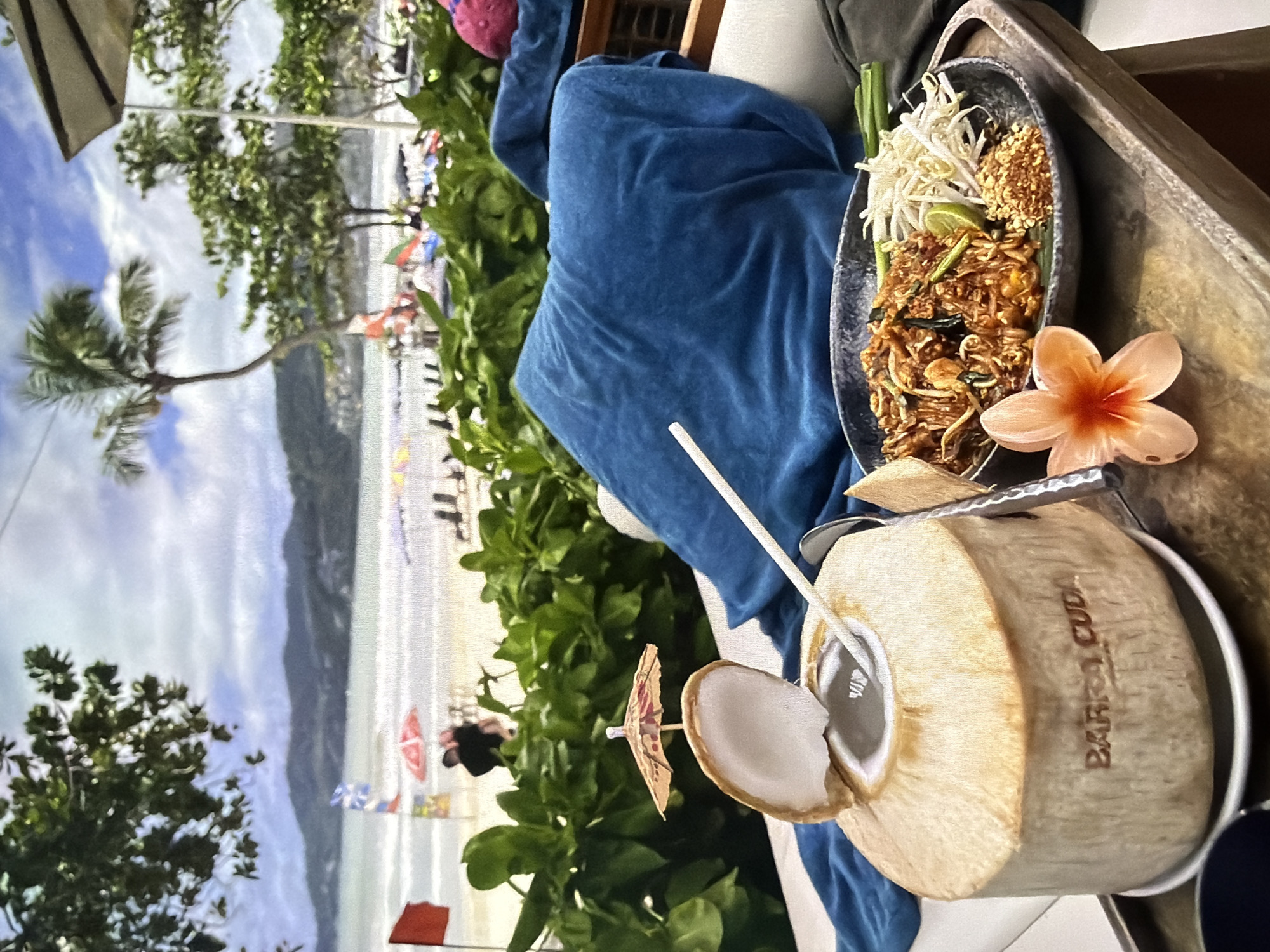 The image displays a vibrant outdoor scene from a first-person perspective, likely at a beachfront restaurant or cafe. The main subjects are a plate of food and a fresh coconut drink, placed on a wooden table.
The food appears to be Pad Thai, served in a dark, textured bowl. It features flat noodles, shrimp or other protein, and is garnished with fresh bean sprouts, crushed peanuts, a lime wedge, and green onions. Beside the plate, a light orange plumeria (frangipani) flower adds a tropical touch.
To the right, a young, husked coconut serves as a drink, complete with a white straw and a miniature paper umbrella. The word "BARRACUDA" is embossed or carved onto the side of the coconut. A metal spoon or small knife is also visible near the coconut.
In the background, a beautiful tropical beach stretches out, featuring light sand, turquoise ocean waters, and distant mountains under a bright, cloudy sky. Palm trees and lush green foliage frame the view. Several beach umbrellas and what appear to be people are visible on the sand, suggesting a popular tourist destination. A blue velvety towel or garment is draped over a chair or railing in the foreground, next to the table.
The scene depicts a relaxing meal or snack during daytime, possibly late morning or early afternoon given the bright natural light. The presence of Pad Thai, a fresh coconut, and tropical foliage strongly suggests a Southeast Asian location, likely Thailand. The overall atmosphere evokes a sense of vacation and leisure.