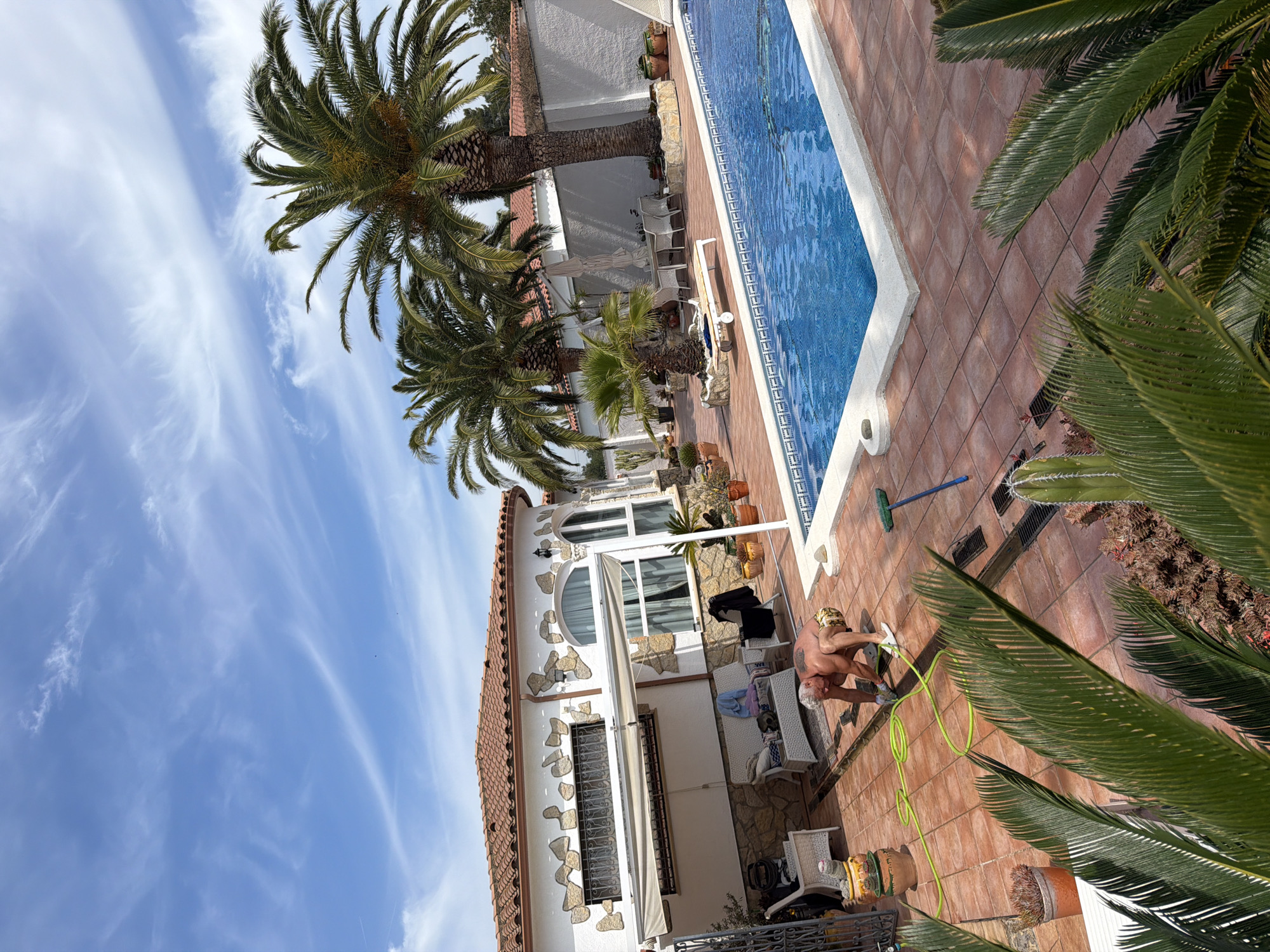 The scene is an outdoor view of a residential area in Mont-roig del Camp, Spain, capturing a sunny day beside a swimming pool. Palm trees punctuate the scene against a sky streaked with wispy clouds. A white, two-story house with a terracotta tile roof stands adjacent to the pool area. A man wearing a patterned head covering is scrubbing the ground near the pool with a scrub brush and a green hose. The pool itself features blue water and a white tiled edge. Potted plants are scattered around the pool area. The scene is captured with a wide-angle lens, introducing some distortion.