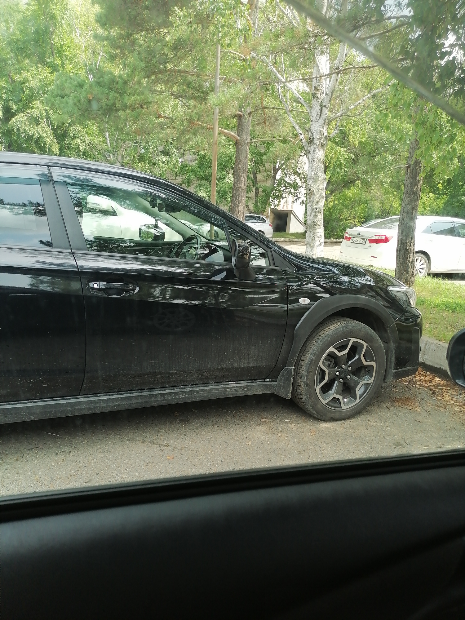 A black Subaru Outback is parked on a gravel and asphalt surface. The car is viewed from the side, with the front passenger side wheel prominently displayed. The car is dusty, with streaks of dirt visible on the doors and body panels. Behind the Subaru, a white Toyota Camry is parked, and to the left of the Subaru, a white structure with a dark entrance is visible. The scene is set in an outdoor environment with trees and foliage providing shade. The image appears to have been taken through a car window, as evidenced by the dark frame at the bottom of the image. This scene is likely located in the city of Lesozavodsk, Russia.