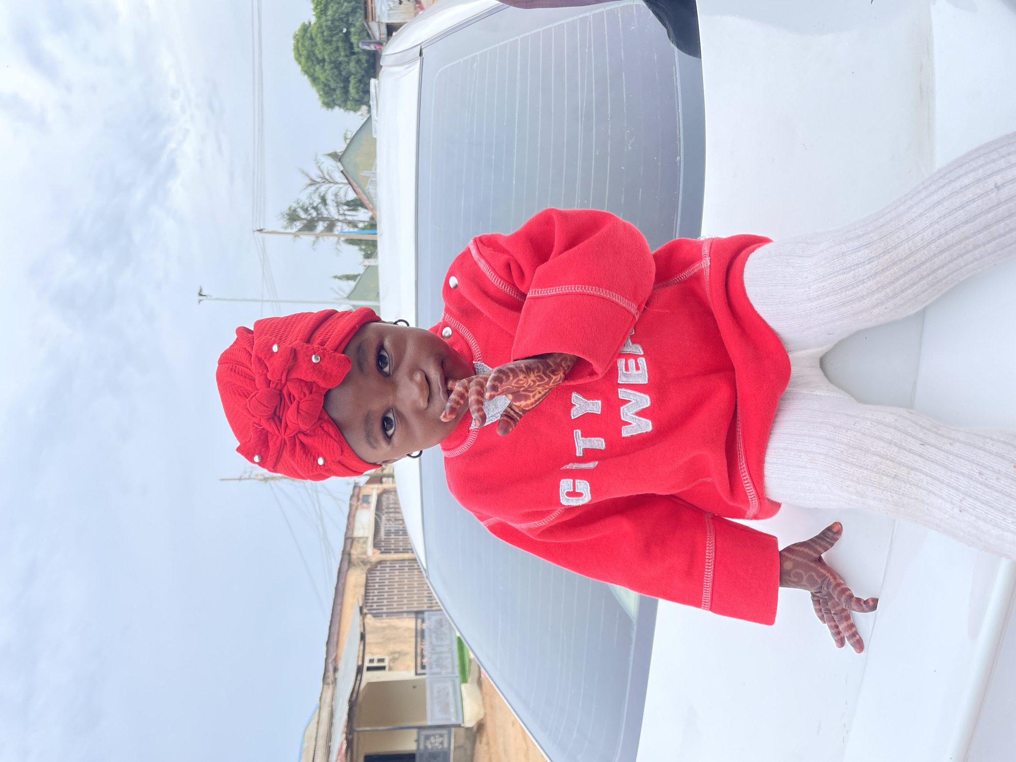 A young child with dark skin is sitting on a white vehicle outdoors in Barakin Akawo, Nigeria. She is wearing a red headscarf with decorative accents, a matching red sweatshirt with the words "CITY WEAR" in white lettering, and white ribbed leggings. Her hands are adorned with henna designs. The background reveals a mix of residential buildings and vegetation under a cloudy sky.