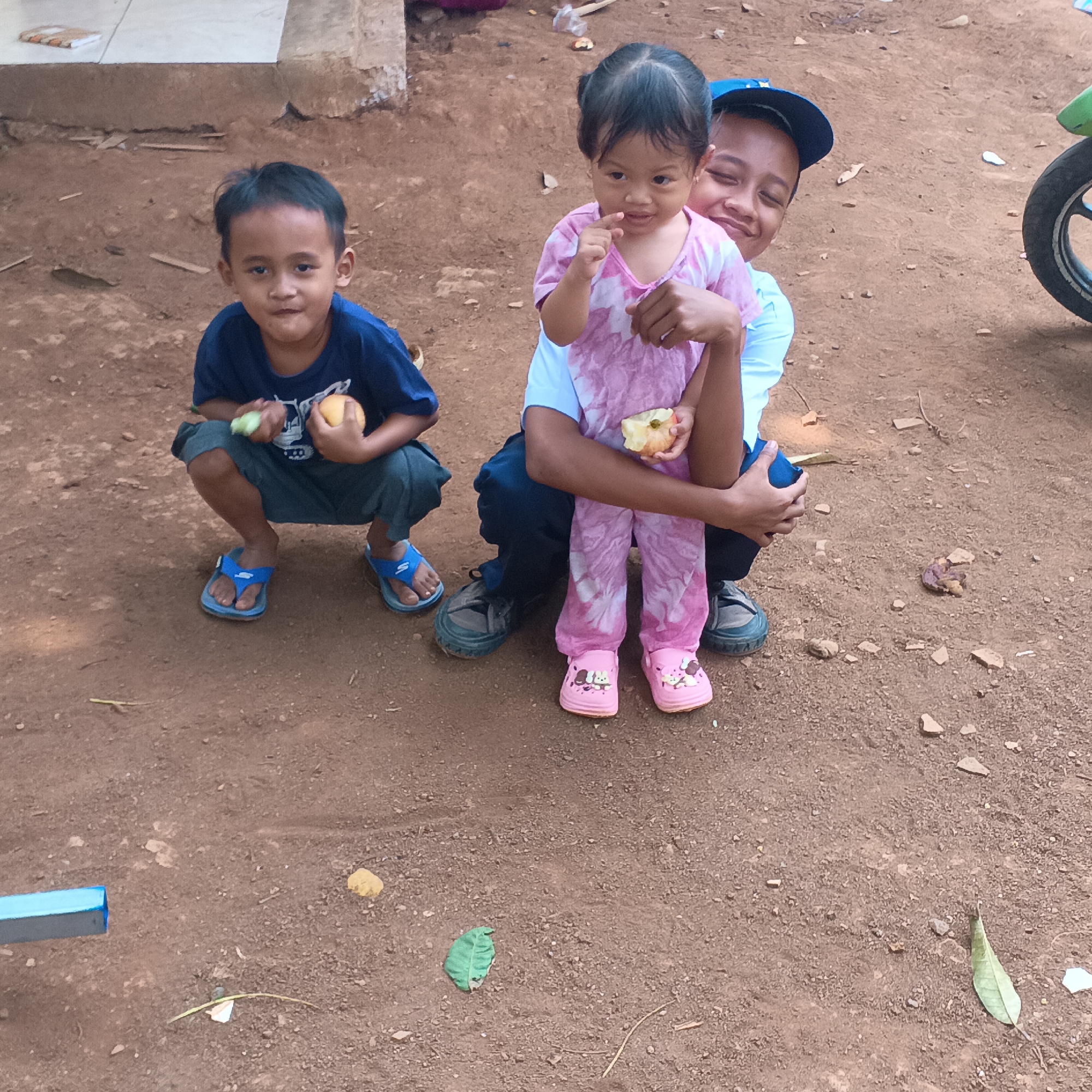 In the city of Jayasampurna, Indonesia, three children are seen outdoors on a dirt ground. The leftmost child, a boy, is crouched down, wearing a blue t-shirt and dark shorts with blue flip-flops. He is holding a light-colored fruit in his left hand and a green vegetable in his right. Beside him, a younger girl is being held by an older boy. The girl, wearing a pink tie-dye top and light-colored pants with pink cartoon-themed shoes, has her finger to her mouth and is holding a piece of fruit. The older boy, wearing a light blue shirt and a blue cap, is behind the girl, with his eyes closed and a smile on his face. In the background to the right, the rear wheel of a motorcycle is visible. The ground is covered with dirt, small stones, and a few fallen leaves. A blue metal object is partially visible in the lower left corner.