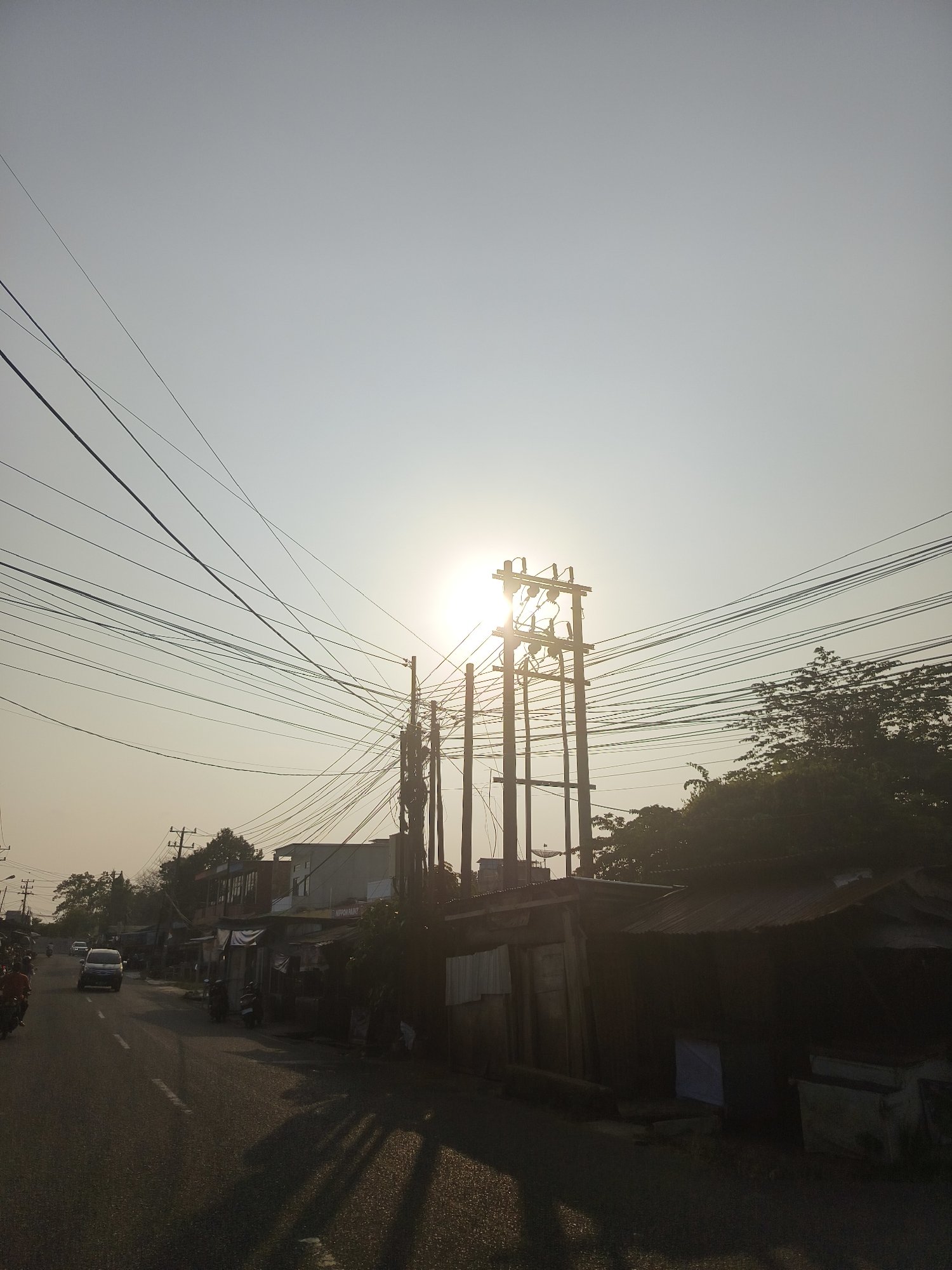 The image depicts a street scene in Pangkalpinang, Indonesia, at what appears to be sunrise or sunset, given the low angle of the sun. A paved road runs diagonally from the lower left to the upper right of the frame. Several vehicles, including a silver car and motorcycles, are present on the road. The left side of the street is lined with simple buildings and structures, some with corrugated metal roofs, and numerous utility poles with a dense network of electrical wires crisscrossing the sky. The sun is positioned behind a utility pole in the upper center of the image, creating a lens flare effect and silhouetting the pole and its components. Trees and vegetation are visible on the right side of the street and behind some of the buildings. Shadows are cast across the road, indicating the time of day. The sky is clear and hazy, with the sun's brightness dominating the upper portion of the image.