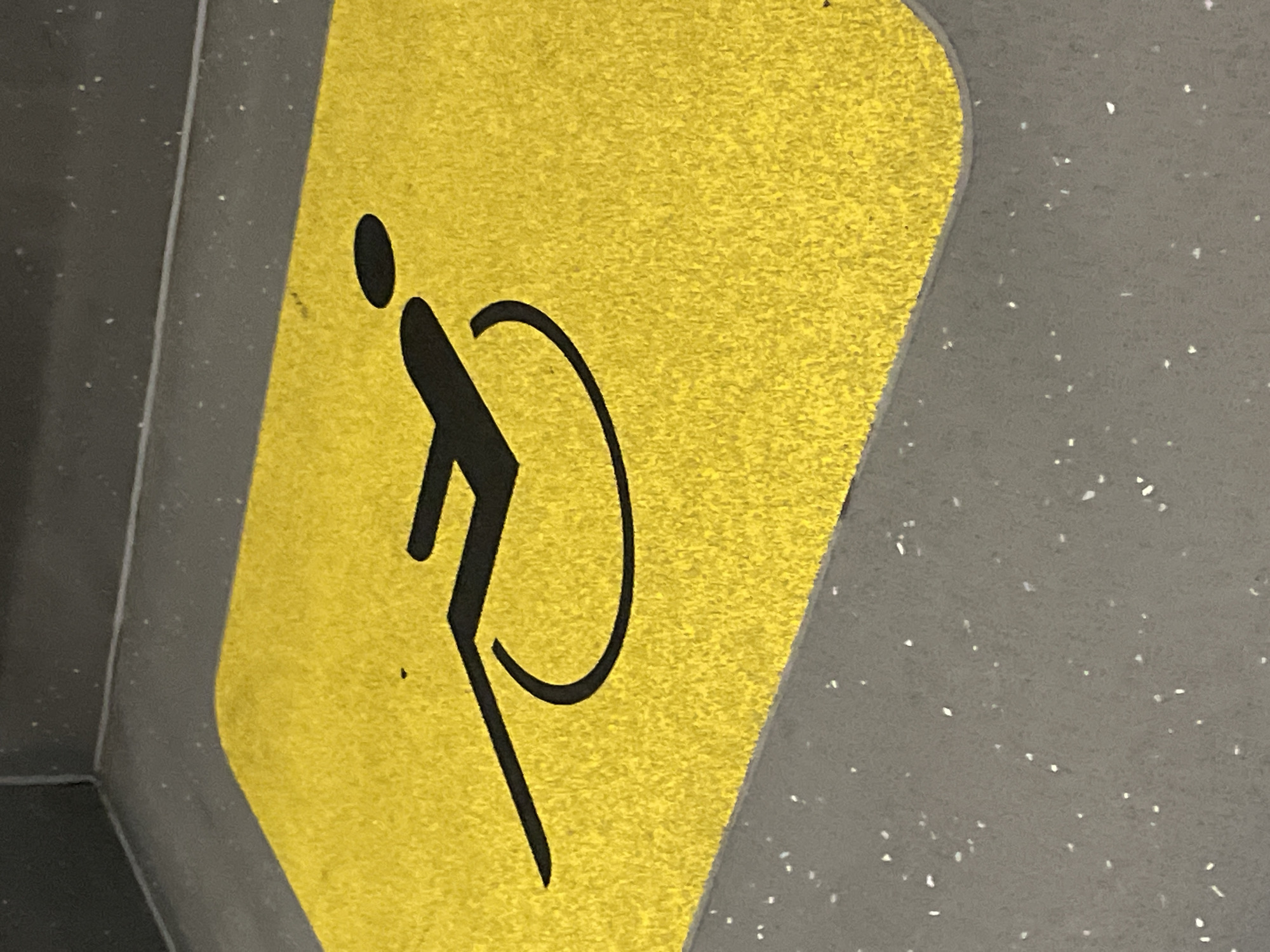 The image shows a handicap sign in Barcelona, Spain. The sign, a bright yellow square, is adhered to a grey speckled surface, possibly flooring. A stylized black figure, commonly used to denote accessibility for people with disabilities, is prominently displayed on the yellow background. The figure is composed of simple lines and shapes, depicting a person leaning forward.