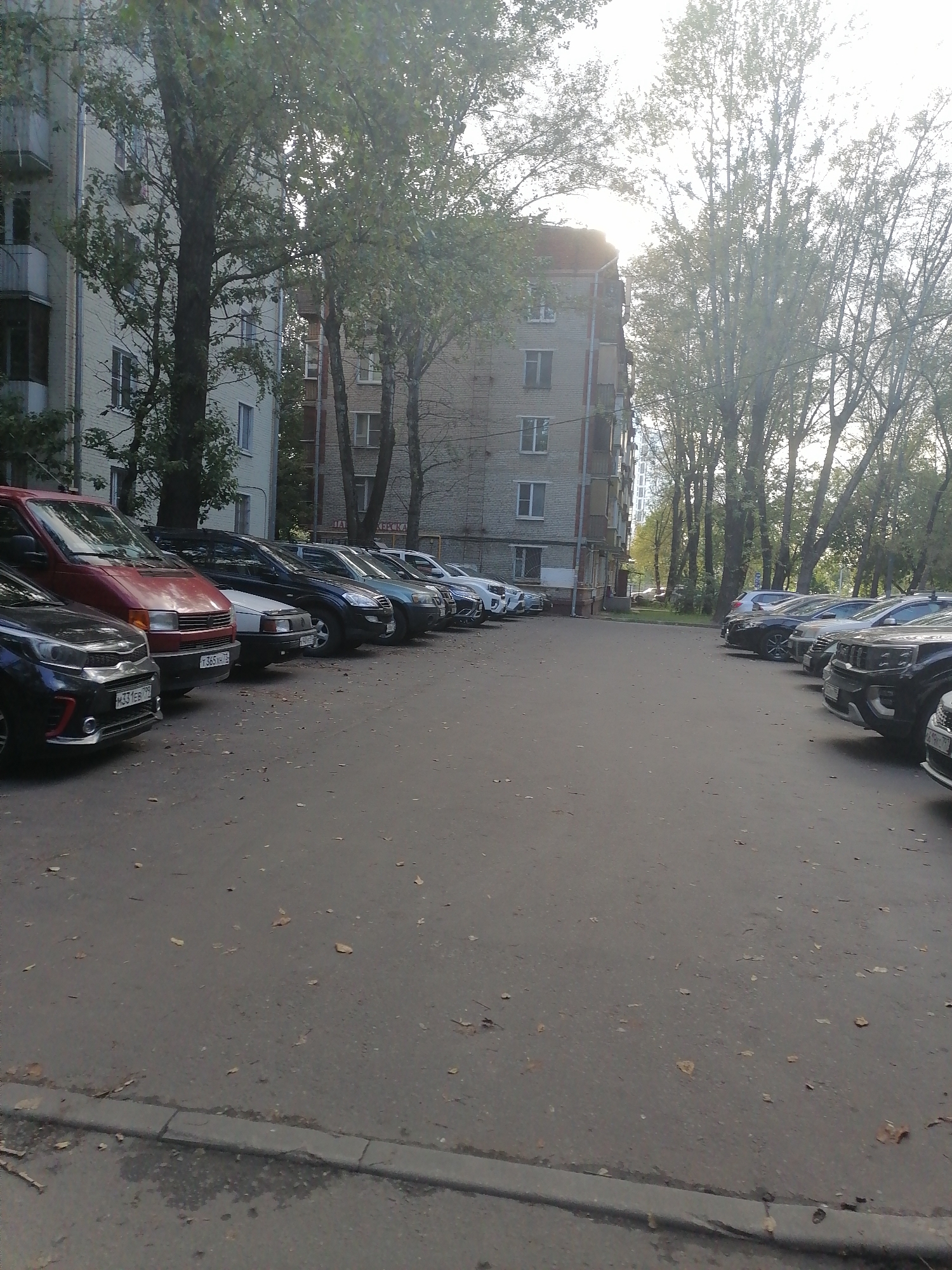 The image depicts a street scene with multiple parked cars and apartment buildings in Moscow, Russia. The foreground is dominated by a paved road with fallen leaves scattered across its surface. Several cars are parked in a line on the right side of the image, while a red minivan and a dark-colored car are parked on the left. The buildings in the background appear to be residential structures, typical of urban Moscow. Trees with green foliage line the street, suggesting it is springtime or summer. The lighting indicates it is daytime, with the sun partially obscured by trees and buildings, casting shadows. The overall impression is one of a typical urban environment in Moscow, with everyday activities like parking and the presence of residential buildings.