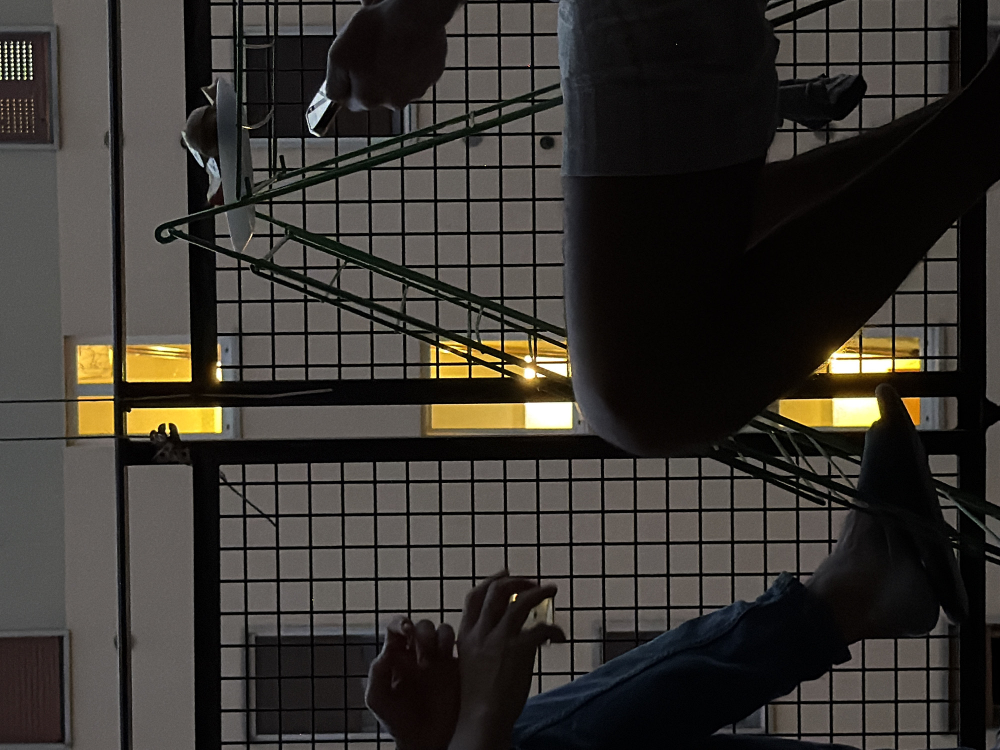 The image shows a human hanging laundry on a clothesline rack on a balcony. The location appears to be in Milan, Italy. A person's legs and torso are visible as they lean over the railing. The person is wearing blue denim pants and possibly shorts, and using both hands. Visible behind the balcony is a grid-like metal structure and a building facade. Windows are visible with warm light emanating from inside.
