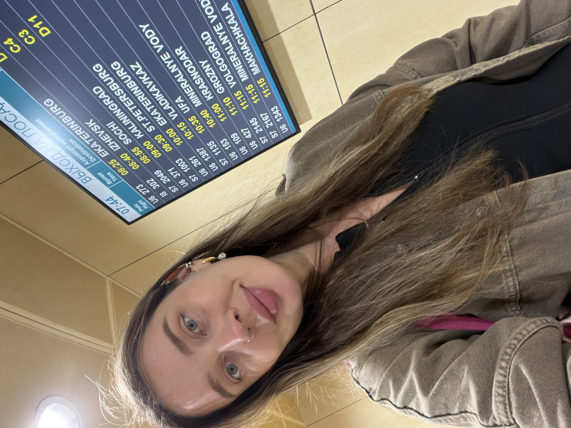 The image shows a woman with long brown hair, wearing a black top and a light brown denim jacket, taking a selfie at Domodedovo Airport in Russia. Behind her is a flight information display board. The board lists various destinations, including Ekaterinburg, Izhevsk, Sochi, Kaliningrad, St. Petersburg, Vladikavkaz, Ufa, Grozny, Krasnodar, Mineralnye Vody, Makhachkala, and Volgograd, along with their flight times and flight numbers. The departure gate information for Ekaterinburg is C4, for Izhevsk is C3, and for St. Petersburg is D11. The time is 07:44. The woman is in focus, and the background is slightly blurred. The lighting appears to be from indoor airport lighting.
