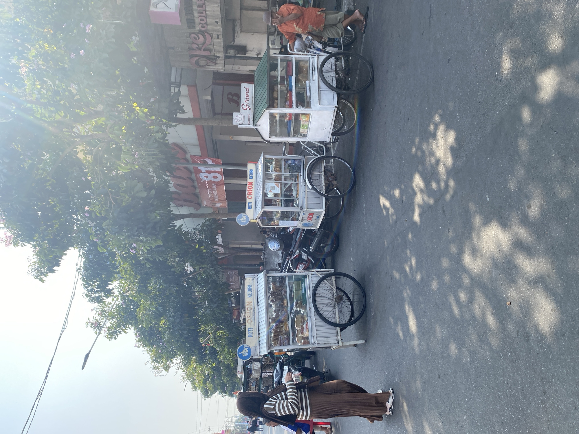 In Kediri City, Indonesia, a street scene unfolds with two food carts selling baked goods. The cart on the left displays "ROTI ORION KEDIRI" and is filled with various loaves of bread and pastries. To its right, another cart, adorned with a green roof and the sign "Grand BAKERY," also showcases an assortment of baked goods. A woman wearing a hijab and a brown striped shirt and skirt walks past the first cart. In the background, a man in an orange shirt and shorts stands near the second cart, appearing to be interacting with it. Motorcycles are parked behind the carts. The environment suggests a sunny day on a paved street lined with buildings and trees, casting shadows on the road.