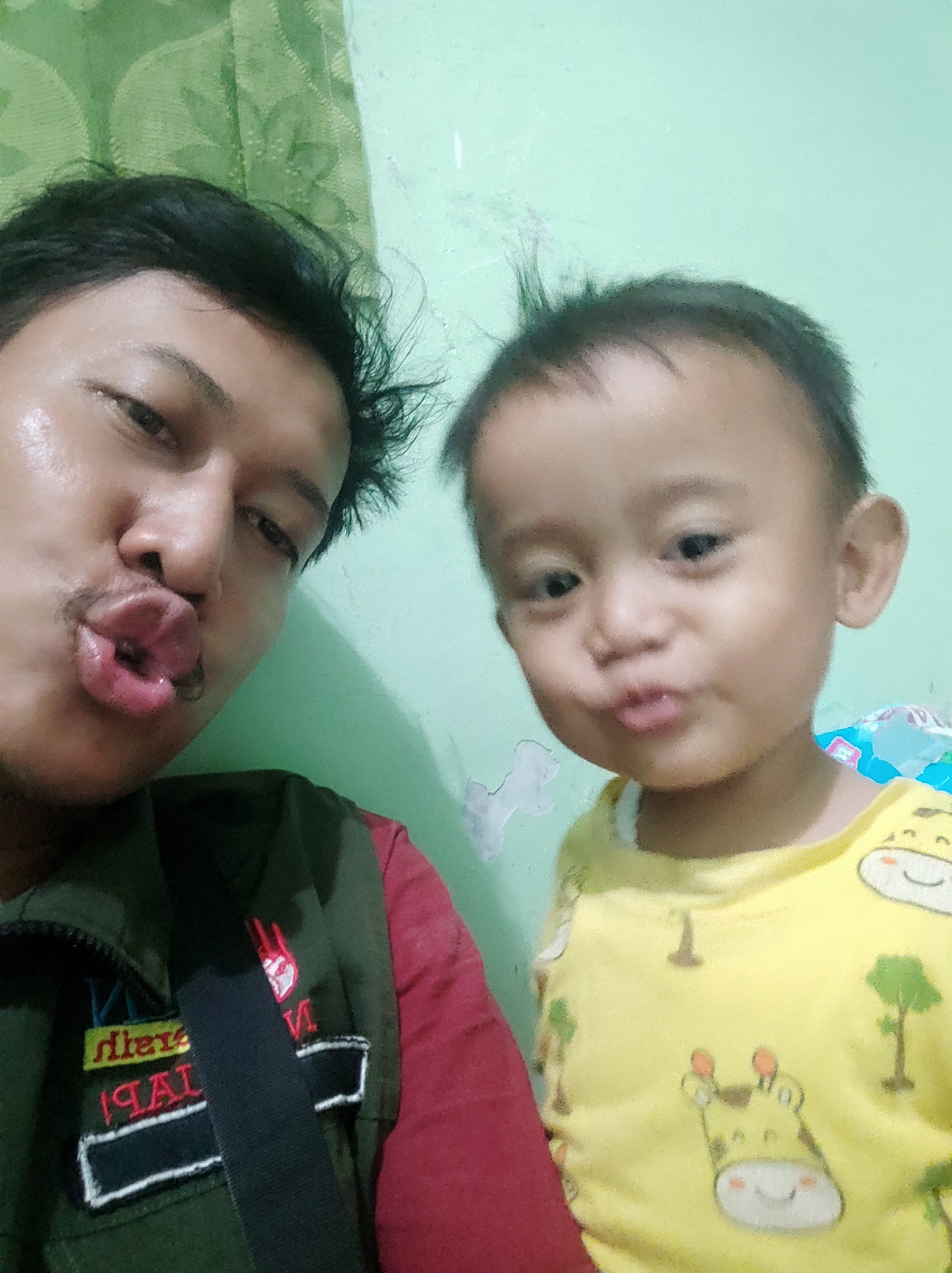 A selfie captures two individuals, an adult male and a young child, in a close-up shot. The adult, positioned on the left, puckers his lips, making a kissing gesture towards the camera. He is wearing a dark green vest over a red shirt. The child, on the right, mirrors the adult's action with pursed lips. The child is dressed in a yellow t-shirt adorned with cartoon giraffes and trees. The background appears to be an interior wall, painted a light mint green, with a section of green patterned fabric, possibly a curtain or tapestry, visible on the upper left. The lighting suggests an indoor setting. This image was taken in Gebang, Indonesia.