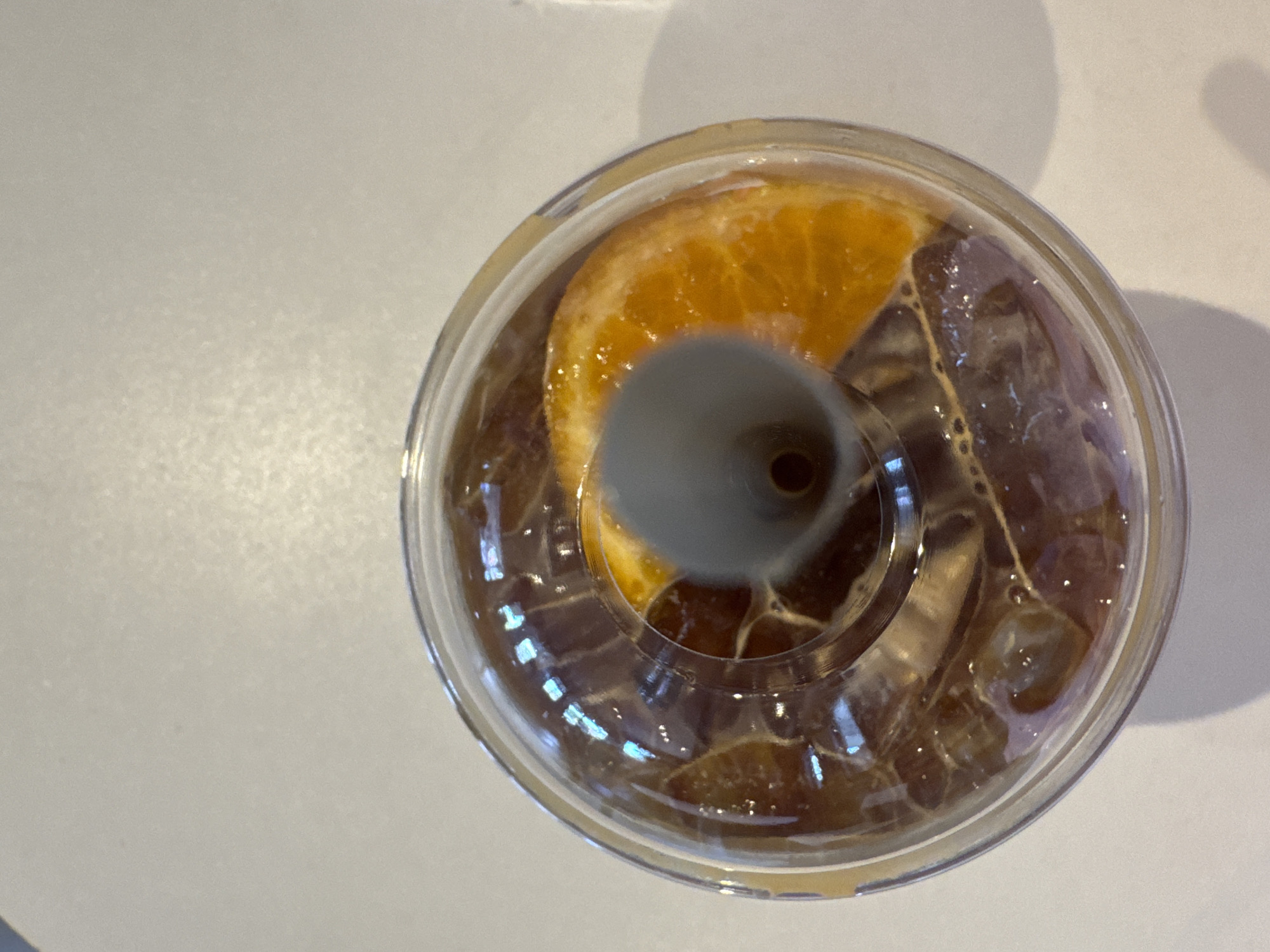 An overhead shot shows a clear plastic cup filled with iced coffee and a slice of orange. The coffee is dark brown and appears to be cold, with ice cubes visible beneath the surface. A gray plastic straw is inserted into the center of the lid. The cup sits on a white surface, possibly a table, with a subtle shadow cast to the right. The scene is indoors, with soft lighting. No people are visible in the image. The location is described as Surat Thani City Municipality, Thailand.