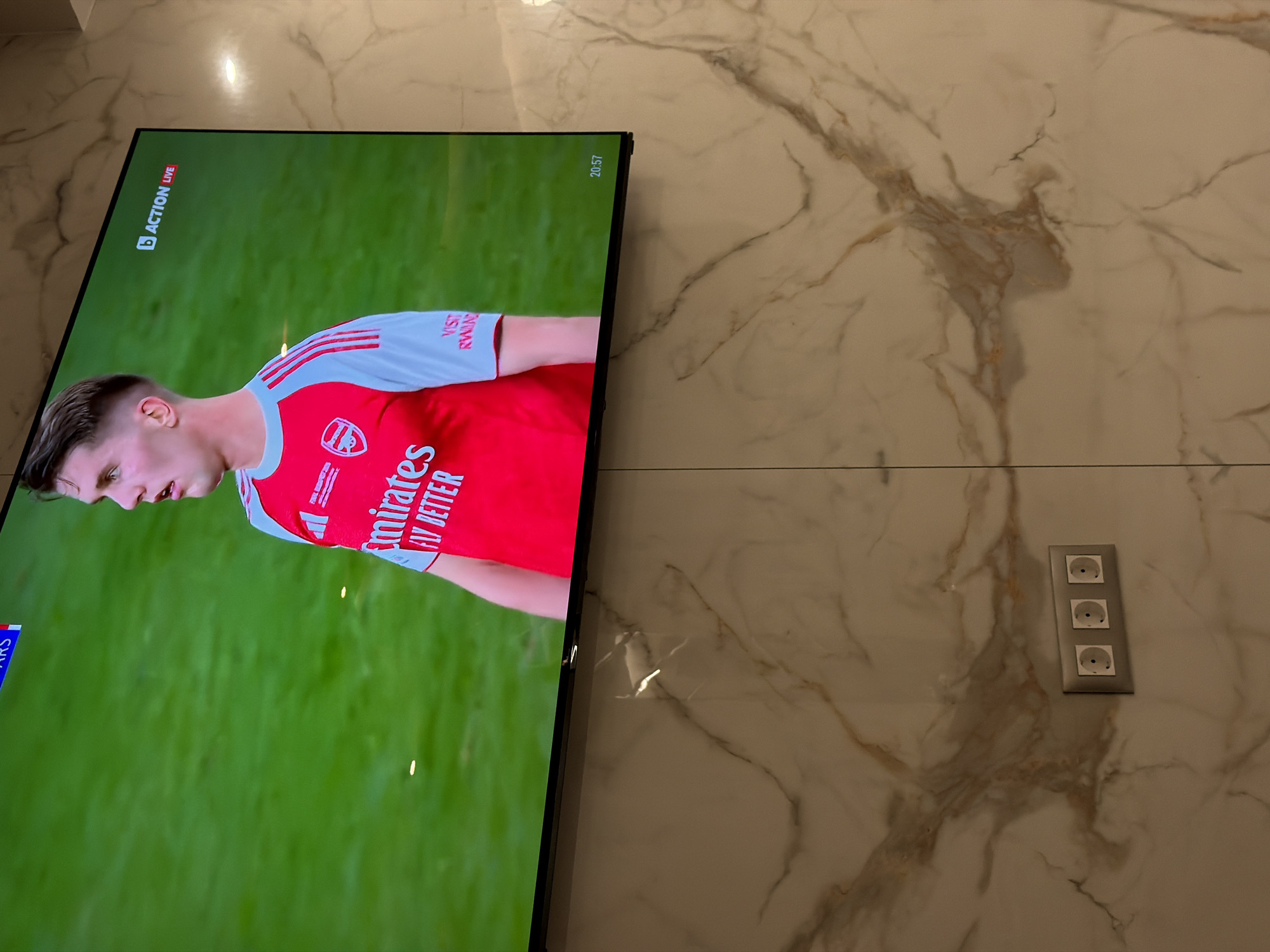 The image shows a television displaying a live broadcast of a soccer game, featuring a player in an Arsenal FC jersey. The broadcast includes "ACTION LIVE" and a time stamp of "20:57". The television is mounted on a marble-like wall, with a triple electrical socket visible on the wall to the right of the screen. The location appears to be indoors, but the specific location in the city of Asenovgrad, Bulgaria, cannot be determined from the image.