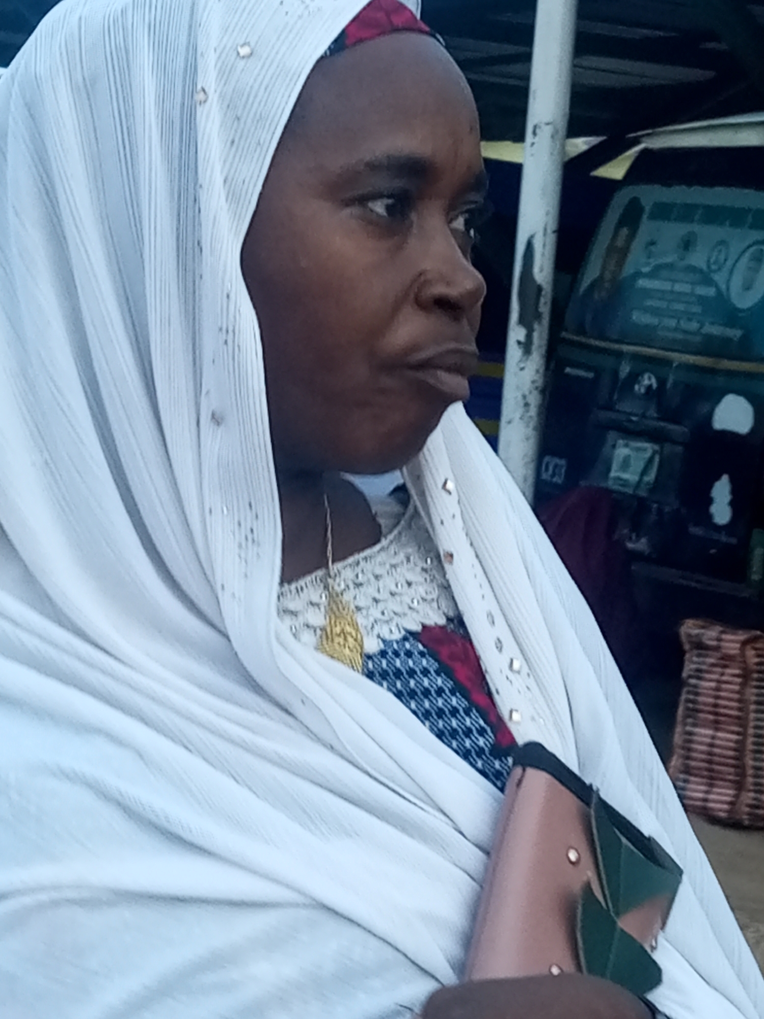 A medium shot captures a person wearing a white hijab, a gold pendant necklace, and a patterned dress. The hijab is adorned with small, sparkly embellishments. The person's face is partially visible, with their head turned to the side. A portion of a brown and green clutch bag is held in their hand, and a wicker basket is visible in the background. The scene appears to be outdoors, possibly in a market or by a roadside in Gombe, Nigeria, as suggested by the presence of a vehicle with signage in the background. The overall lighting is natural and subdued.