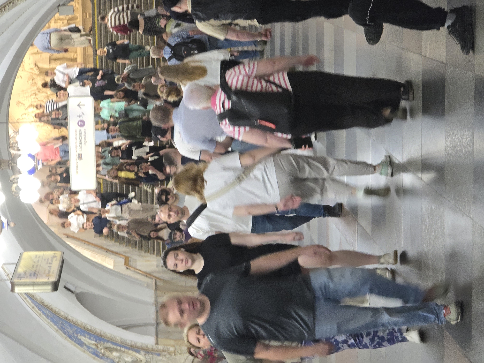 The image captures a crowded scene in a Moscow subway station. Numerous people are walking in both directions on a staircase. The station features arched ceilings, tile mosaics, and a signage indicating directions for Taganskaya station. A large crowd is descending the stairs, while another group is ascending. The color scheme is dominated by muted colors, with various shades of grey, white, and blue. The scene conveys a sense of everyday movement and hustle within the transit infrastructure of Moscow.