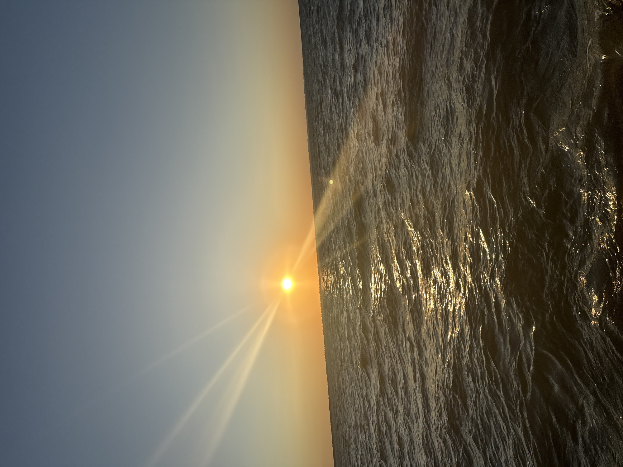 The image depicts a serene sunset over the Black Sea, viewed from the coastline of Zatoka, Ukraine. The sun, a vibrant orange disc, is low on the horizon, casting long, ethereal rays of light across the sky and water. The sky is a gradient of soft blues and oranges, transitioning into a pale yellow near the sun. The sea is calm, with gentle ripples reflecting the golden light. A faint line of distant buildings or structures can be seen along the horizon, suggesting the presence of the town of Zatoka. No people or specific objects are identifiable in the scene, focusing the viewer's attention on the natural beauty of the sunset.