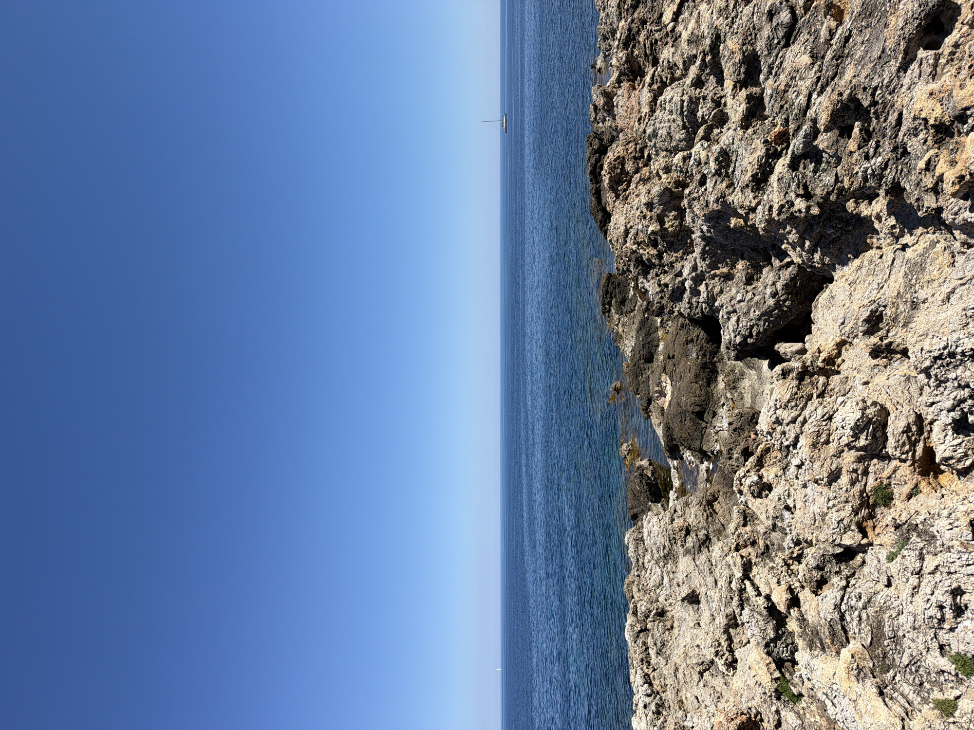 The image depicts a coastal scene in Capdepera, Spain. On the right side of the image is a rocky cliff face, characterized by its rugged texture and variations in color from light beige to dark brown. The sea occupies the central portion of the image, its blue surface rippled. A single sailboat is visible on the horizon. The sky is clear, transitioning from a deeper blue at the top to a lighter shade near the horizon.