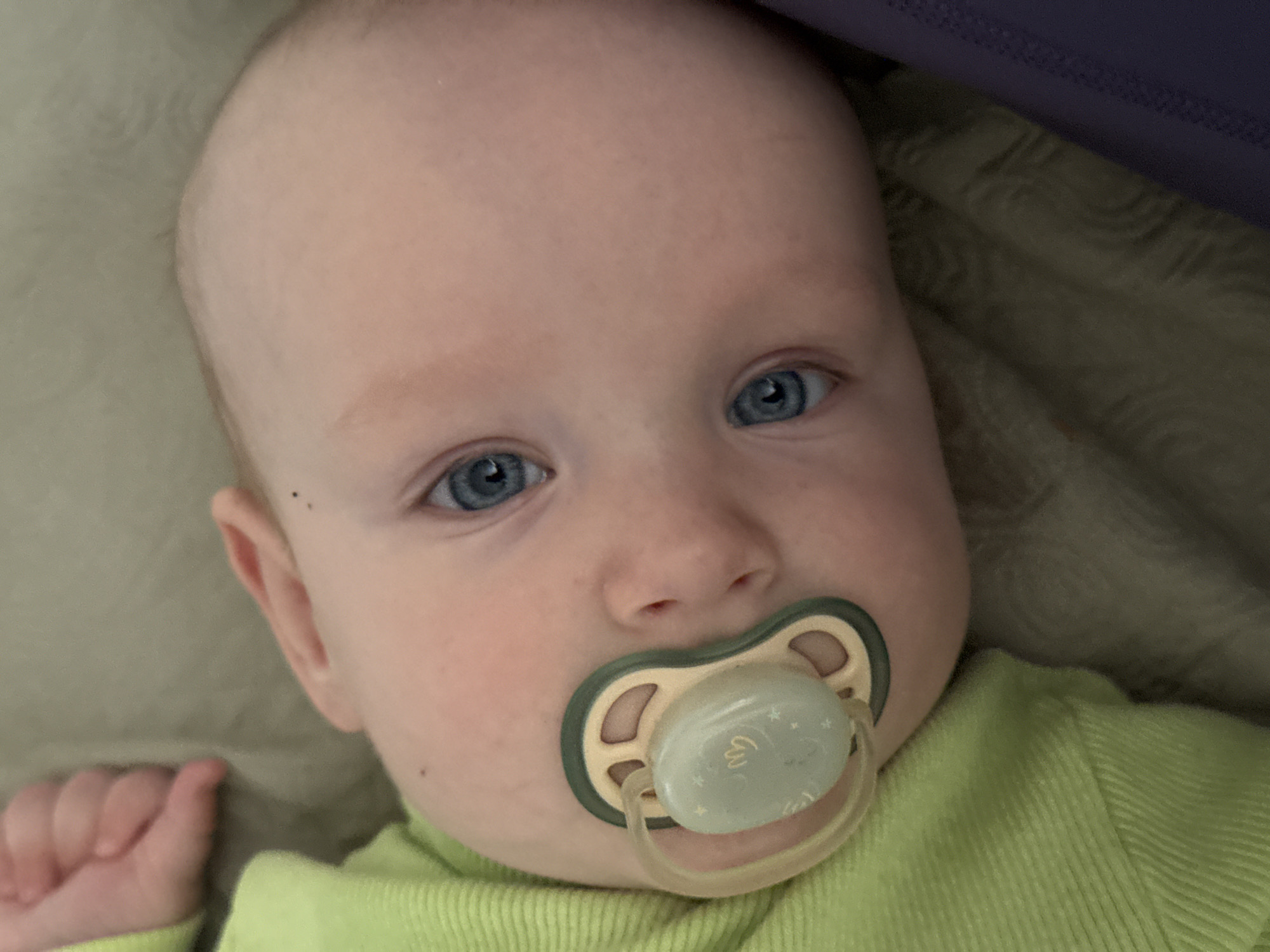 The image shows a close-up, head-and-shoulders shot of a baby with blue eyes and a pacifier in its mouth. The baby is wearing a light green, ribbed garment. The background appears to be a neutral-colored, textured surface, possibly a blanket or couch. The baby's left hand is visible in the upper left corner, appearing to be resting. There is no indication of the city of Moscow, Russia, in the image.
