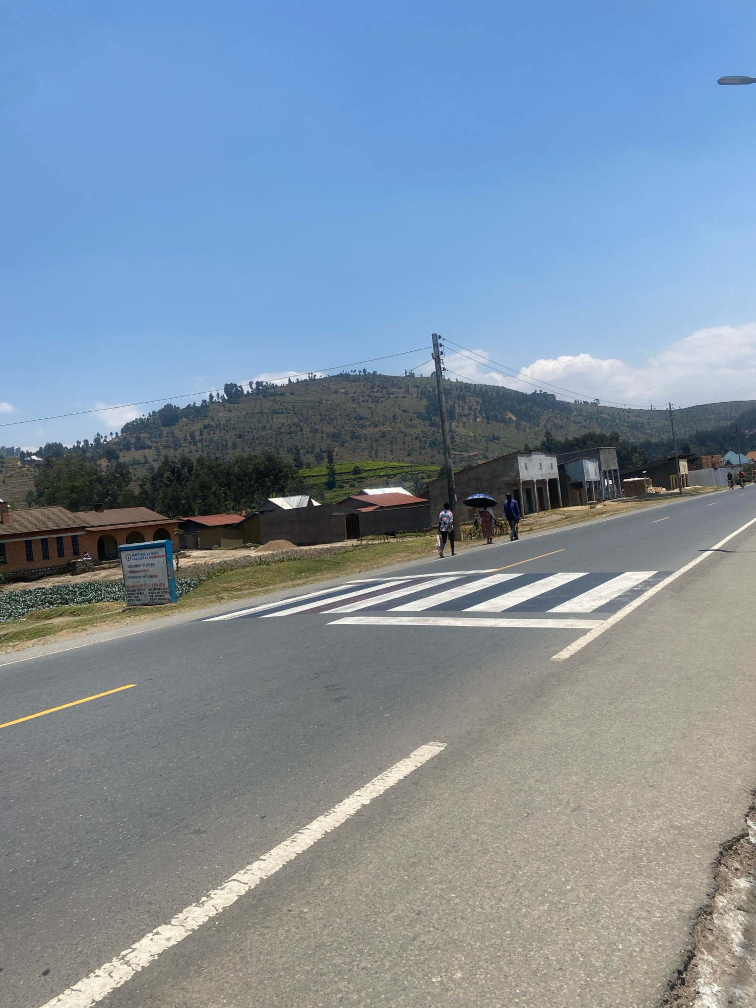 In the city of Shaba, Rwanda, a paved road stretches into the distance under a clear blue sky. A pedestrian crossing, marked with bold white stripes, dominates the foreground. To the left of the road, a brick building with arched windows stands next to a field of green crops. Further down the road, several small, simple structures line the right side, some with corrugated metal roofs. In the middle ground, three people walk across the pedestrian crossing, one holding a blue umbrella. Beyond the buildings, a lush, green hillside rises steeply, dotted with trees and scattered houses. The scene suggests a rural or semi-urban environment with everyday life unfolding along the roadside.