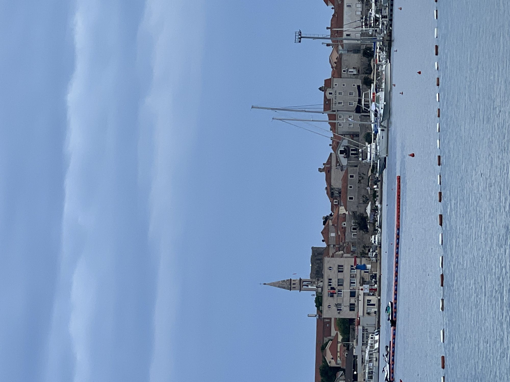The image shows a waterfront view of a European city, likely taken from across a bay or harbor. Buildings with red-tiled roofs and light-colored facades line the shore, interspersed with masts of sailboats and yachts docked in the water. In the foreground, floats mark the boundary of a swimming area. A church steeple rises above the buildings. The sky is a clear blue with some faint streaks of clouds. The city is in Budva, Montenegro.