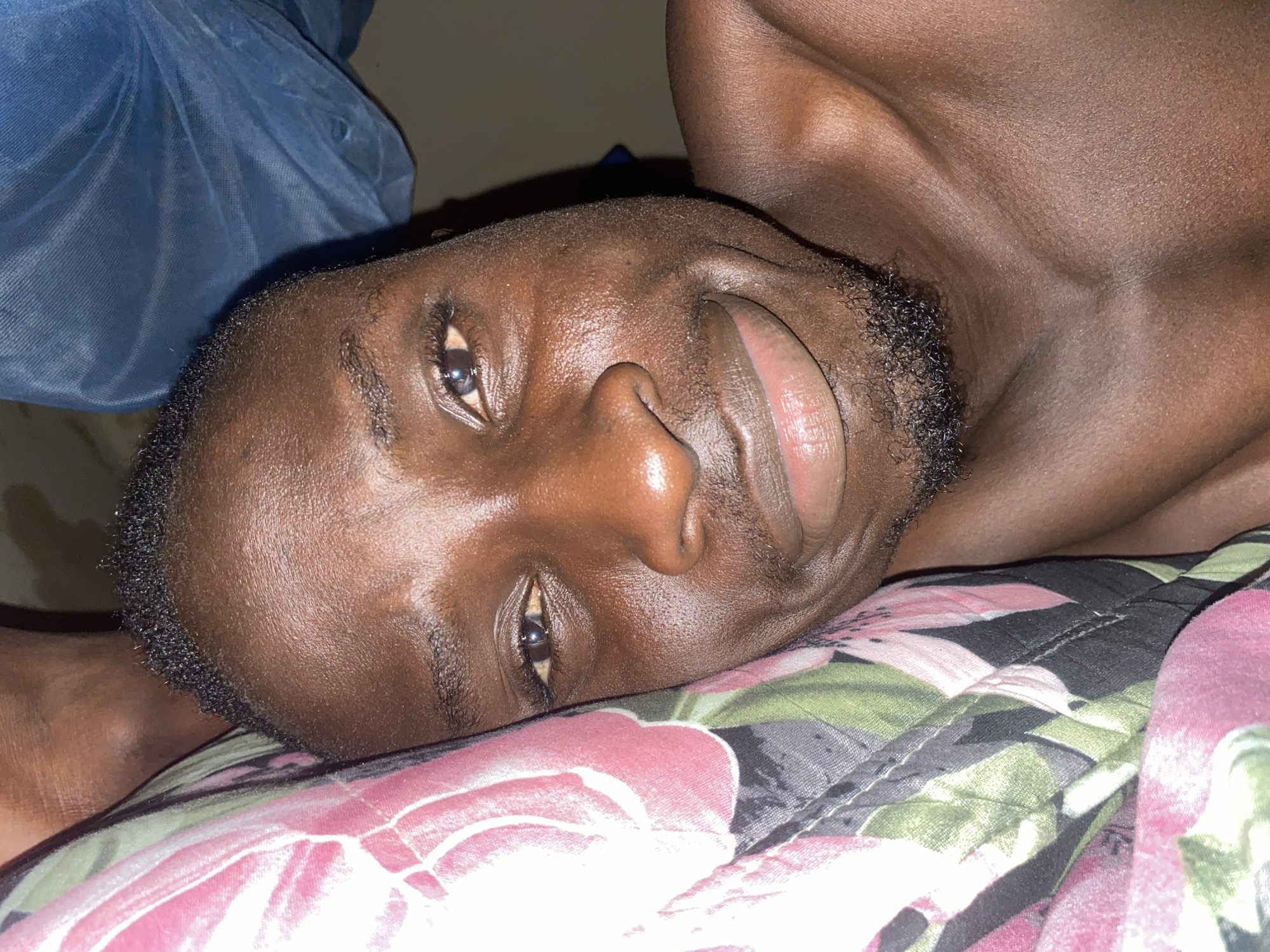 The image shows a close-up, overhead view of a Black person lying on their back. The person's head is tilted to the right, and their eyes are open, looking towards the camera. They have dark skin, short black hair, and a light beard. The person is wearing a blue garment that is partially visible in the upper left corner, and lying on a floral-patterned blanket. The background is dark and indistinct. The overall lighting suggests the image was taken indoors, possibly in a bedroom. No specific location in Barden Yana, Nigeria can be determined from the image.
