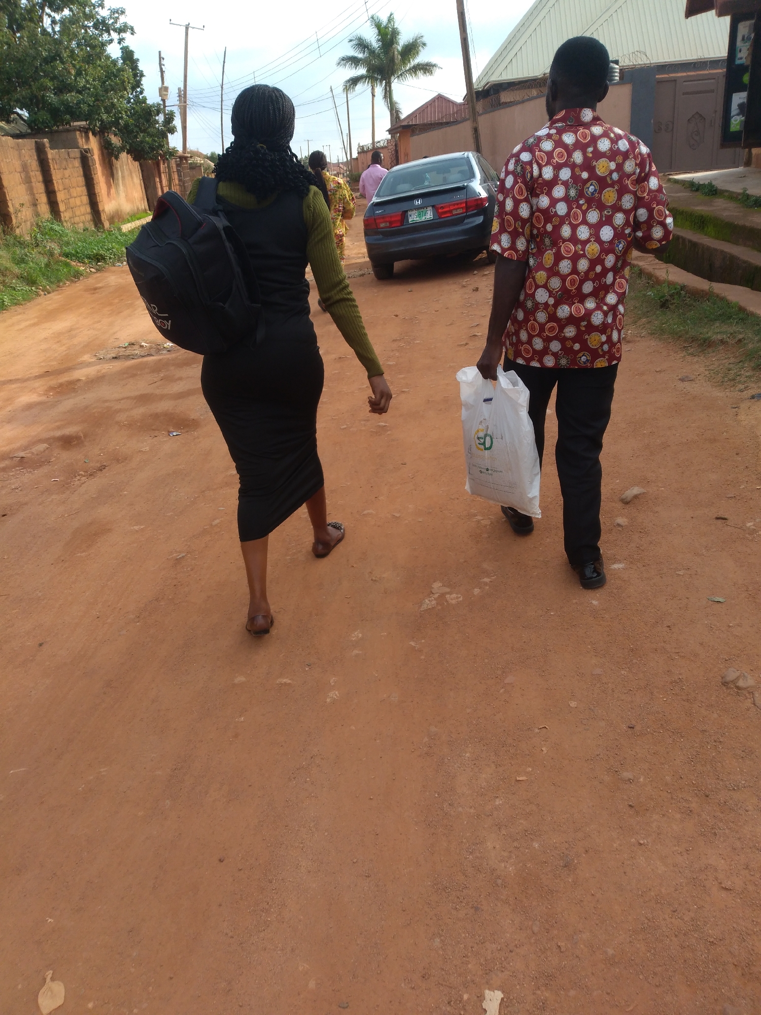 In Barakin Akawo, Nigeria, two individuals are walking away from the camera on a dusty, reddish-brown dirt road. The person on the left is a woman wearing a black backpack, a dark green long-sleeved top, and a black pencil skirt. She has braided hair and is wearing flat sandals. The person on the right is a man wearing a red shirt with a repetitive pattern of pocket watches, black trousers, and black shoes. He is carrying a white plastic shopping bag in his right hand. A dark-colored sedan is parked on the right side of the road behind the individuals. The environment appears to be a semi-urban or rural area, with a brick wall and greenery visible on the left, and buildings and a roof structure on the right. Utility poles with numerous wires are present in the background, indicating the presence of infrastructure. The sky is visible above, with some clouds.