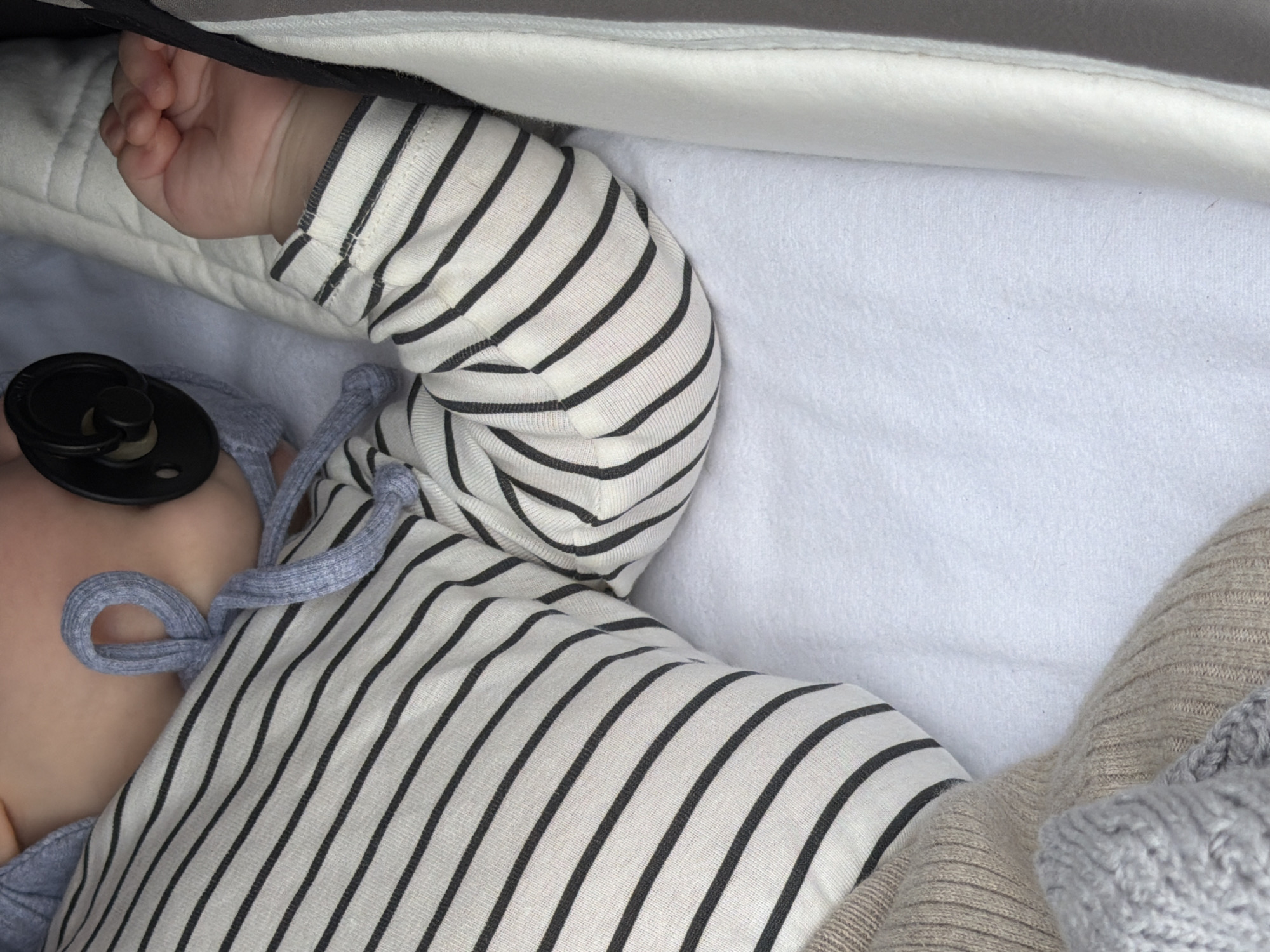 A baby is lying in a white bassinet or stroller. The baby is wearing a striped white and black shirt with long sleeves, and a black pacifier is in its mouth. A light grey blanket is partially visible near the baby's head, and a tan knitted blanket is visible on the right. The baby's hand is clenched into a fist. The top edge of the bassinet/stroller has a grey and white cushioned lining. The location is not discernible from the image provided.
