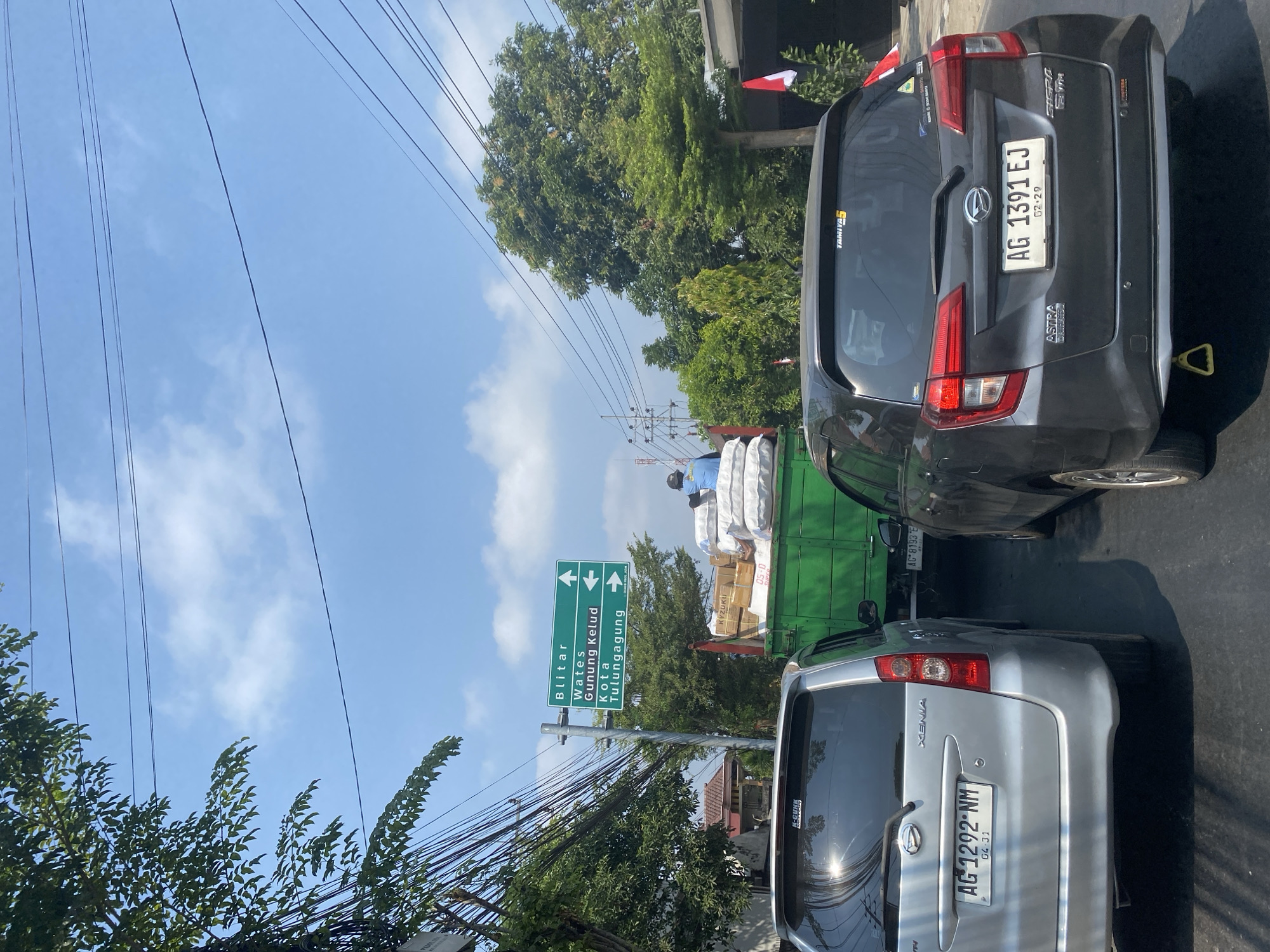 This image captures a traffic scene on a sunny day in Kediri City, Indonesia. In the foreground, two cars are stopped in traffic: a silver Daihatsu Xenia with license plate AG 1292 NM on the left, and a dark grey Daihatsu Sigra with license plate AG 1391 EJ on the right. Behind the cars, a green truck is visible, loaded with sacks of goods and some boxes. A person wearing a blue shirt is standing on the truck. A directional signpost indicates routes to Blitar, Wates, Gunung Kelud, and Kota Tulungagung. The environment includes trees, overhead power lines, and buildings. The sky is blue with scattered white clouds.