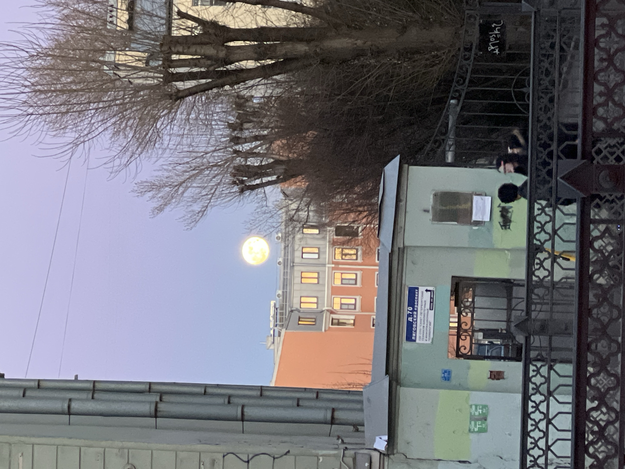 The image depicts a street scene in Saint Petersburg, Russia, with a full moon visible in the sky. The buildings are a mix of old and new, with some featuring orange and gray facades and illuminated windows. A green building with a sign that reads "Лиговский проспект д.70" (Ligovsky Prospekt 70) is situated in the foreground. A black wrought-iron fence runs alongside the building. Bare trees are visible above the buildings. On the right side of the fence, two people are looking at a building or sign.