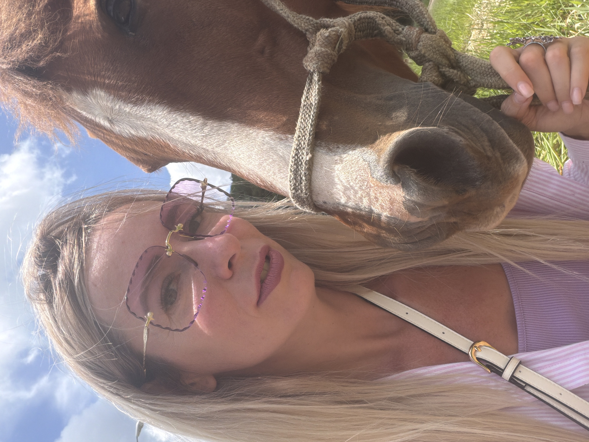 A selfie taken outdoors shows a woman with long blonde hair wearing pink-tinted glasses and a light purple top underneath a pink and white striped jacket. She is holding a rope halter on a brown horse. The horse's head is close to the woman's face, and it appears to be sniffing the camera. The background shows a cloudy sky and green grass, suggesting a rural or natural setting. The location is identified as Da Lat, Vietnam.