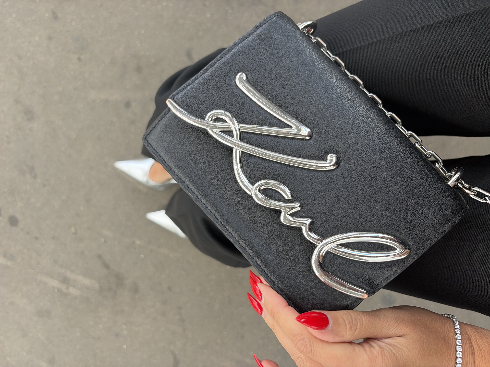A black leather clutch purse with a silver metallic script logo is held by a woman's hand with bright red pointed fingernails. The purse has a silver chain strap. The woman is wearing black pants and pointed silver heels. She also has a diamond tennis bracelet on her wrist. The scene is set outdoors on a grey, textured surface that appears to be pavement.