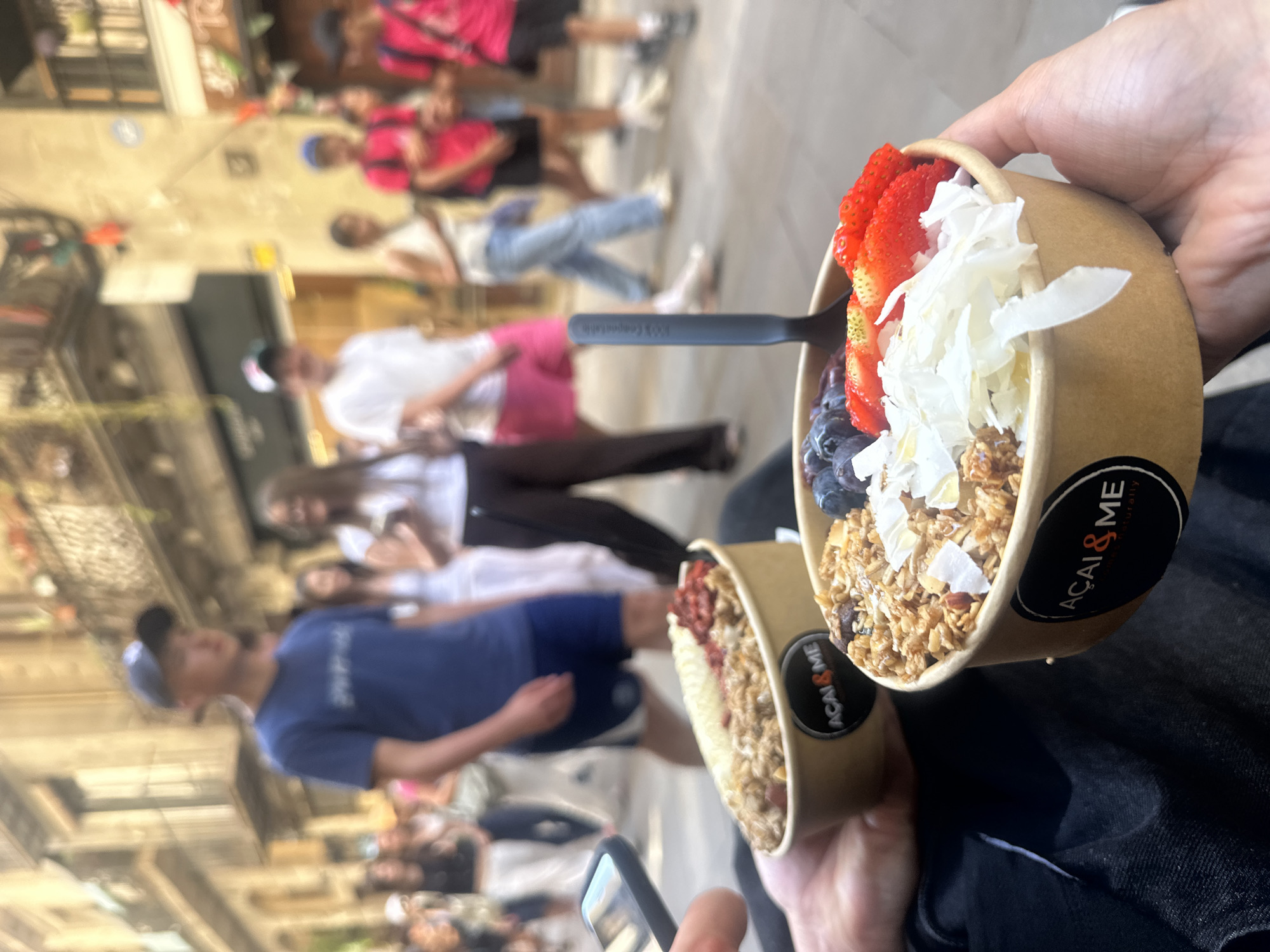Here is a forensic description of the image.
The image shows two paper bowls filled with acai bowls. One bowl contains granola, blueberries, strawberries, and coconut flakes. The other bowl appears to contain granola and strawberries. Each bowl has a black sticker displaying the words "ACAi & ME". A black spoon is inserted into the bowl with more toppings. The bowls are being held in a person's hands.
The background shows a blurry street scene with pedestrians walking by. The setting is an urban environment, potentially Barcelona, Spain.