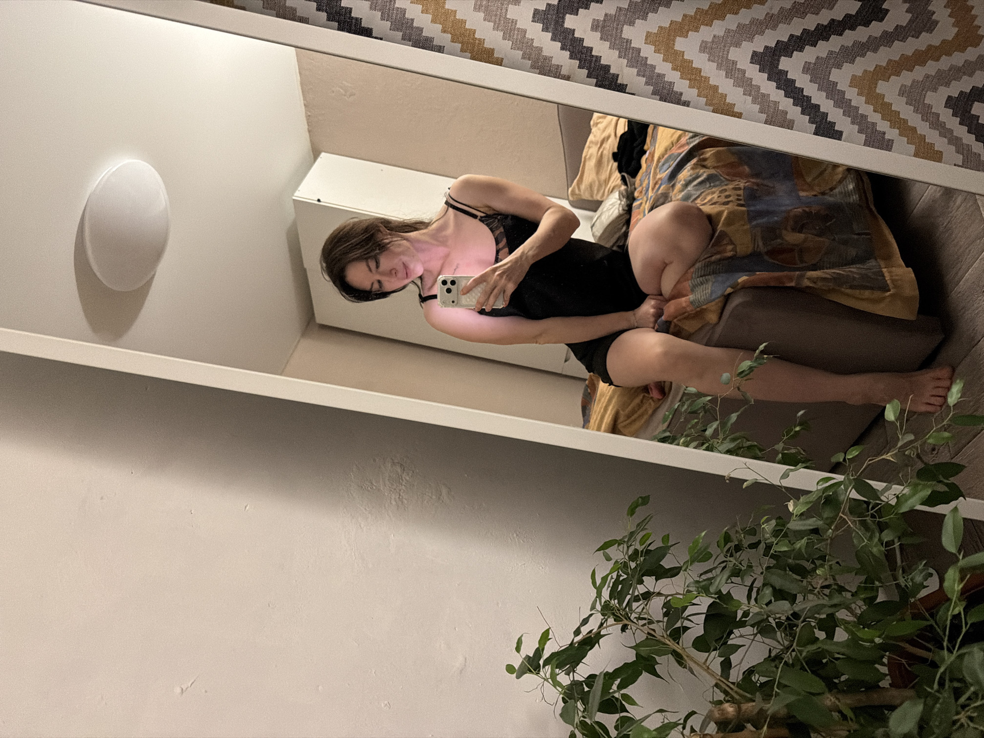 The image depicts a reflection in a mirror. The reflection shows a woman wearing a black garment with straps, holding a smartphone. Behind her is a bed with patterned bedding. To the left of the mirror is a wall with a light fixture. In front of the mirror, a plant is visible. The location is likely a private residence; city and country details are inferred from the prompt, but not verifiable in the image. No action or interactions are visible beyond the woman holding her phone.