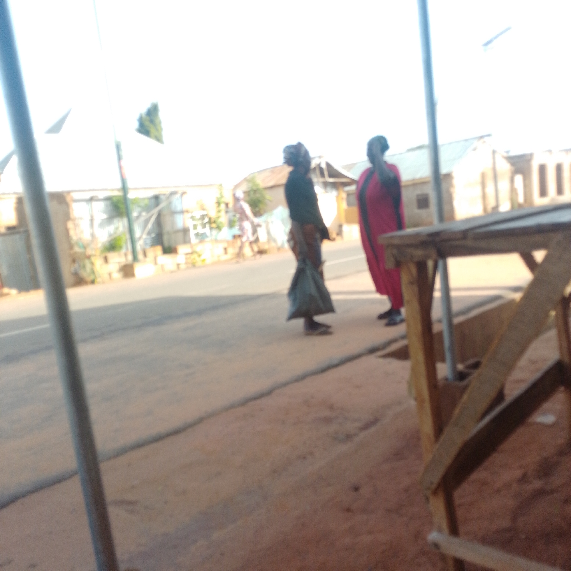 Two people are standing on a dusty street in front of buildings in the city of Tempure, Nigeria. The person on the left is wearing dark clothing and appears to be carrying a dark bag. The person on the right is wearing a bright red outfit. A wooden structure is in the foreground, partially obscuring the view of the street and the individuals. The sky is bright and washed out, indicating daytime. The environment appears to be somewhat rural or underdeveloped.