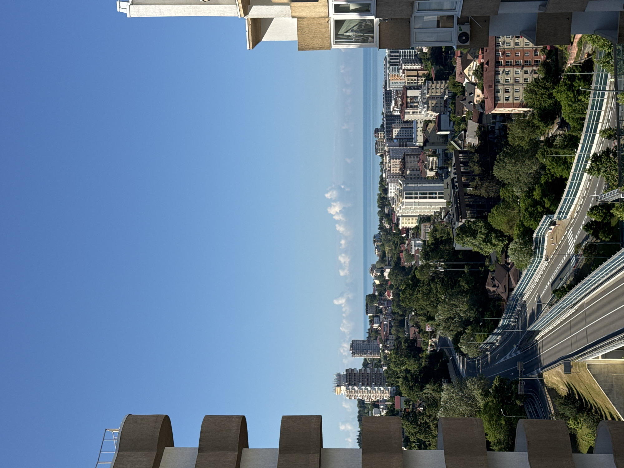 The image shows a view of the city of Sochi, Russia, from a high vantage point. In the foreground, the curved balconies of two modern apartment buildings frame the scene. The buildings are constructed with beige and white concrete.
Beyond the buildings, the city stretches out with a mix of residential and commercial structures, including several tall, modern apartment blocks and some older, lower-rise buildings. Lush green trees and vegetation are abundant throughout the city, suggesting a well-treed urban environment. A multi-lane road winds through the city, with bridges and overpasses. The road is bordered by a white and blue barrier.
In the distance, the calm blue water of the Black Sea is visible, with a clear blue sky overhead. A few scattered white clouds dot the horizon. The lighting suggests it is daytime, likely morning or afternoon, with bright sunlight casting shadows. There are no people visible in the image. The overall impression is one of a vibrant, coastal city nestled amongst greenery, under a clear sky.