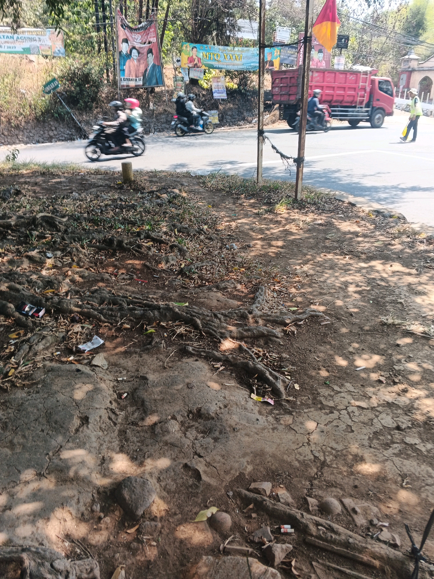The image depicts a street scene in a city that appears to be Semarang, Indonesia, given the context. In the foreground, exposed tree roots and dry leaves litter the cracked, dusty ground, indicating a possibly unkempt or natural area adjacent to the road. A few pieces of trash, including cigarette butts and crumpled paper, are scattered on the ground.
On the road, several vehicles are present. Two motorcycles are moving, one with two riders, and another with a single rider. A large red dump truck is also on the road, suggesting a construction or transport activity. A person in a high-visibility vest, holding a yellow flag, is positioned on the side of the road, possibly directing traffic or involved in an event.
In the background, banners with Indonesian text are visible, some featuring images of people, suggesting local advertisements or campaign materials. The environment is characterized by trees lining the road and a bright, sunny sky, casting shadows on the ground. A small building with an arched entryway is visible in the far background. The overall scene suggests a typical day on a roadside in an Indonesian city, with a mix of daily life, traffic, and local context.