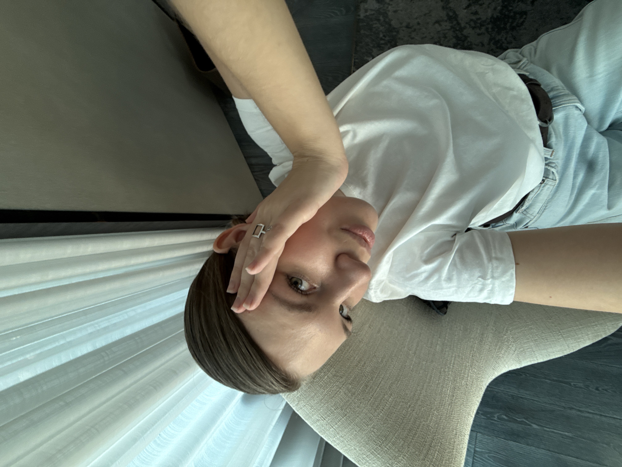 A woman with short, dark brown hair and light skin is photographed in a close-up, tilted perspective. She is wearing a white t-shirt and light blue jeans with a brown belt. Her right hand is raised to her forehead, and a rectangular ring is visible on her finger. The background includes a textured grey chair, a sheer white curtain, and what appears to be a dark grey or black floor. The location is possibly indoors, given the presence of the curtain and chair. The scene's composition is relatively simple, focusing on the woman and her posture, which suggests possible fatigue or concern. It's impossible to definitively determine the location is the city of Vracar Urban Municipality, Serbia, without further contextual information.