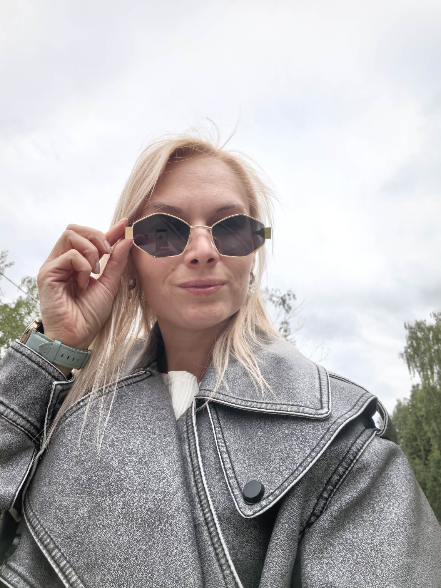 In an outdoor setting, a fair-skinned woman with blonde hair and sunglasses is captured from a low angle. She is wearing a grey leather jacket over a white top and a light blue smartwatch on her left wrist. The woman is holding her right hand up to adjust her sunglasses. The background consists of a cloudy sky and green trees, suggesting a natural or park-like environment. There is no indication of the specific location within Sterlitamak, Russia.