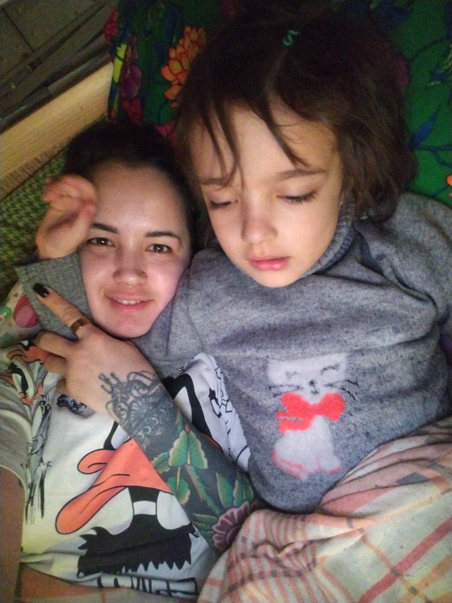 A woman and a child lie together. The woman is smiling, has tattoos on her arm, and a ring on her finger. The child is wearing a grey sweater with a white cat design and a red bow. The woman is wearing a t-shirt with a cartoon duck. They are lying on a patterned surface. The location is not identifiable as Гожан, Russia from the provided image.