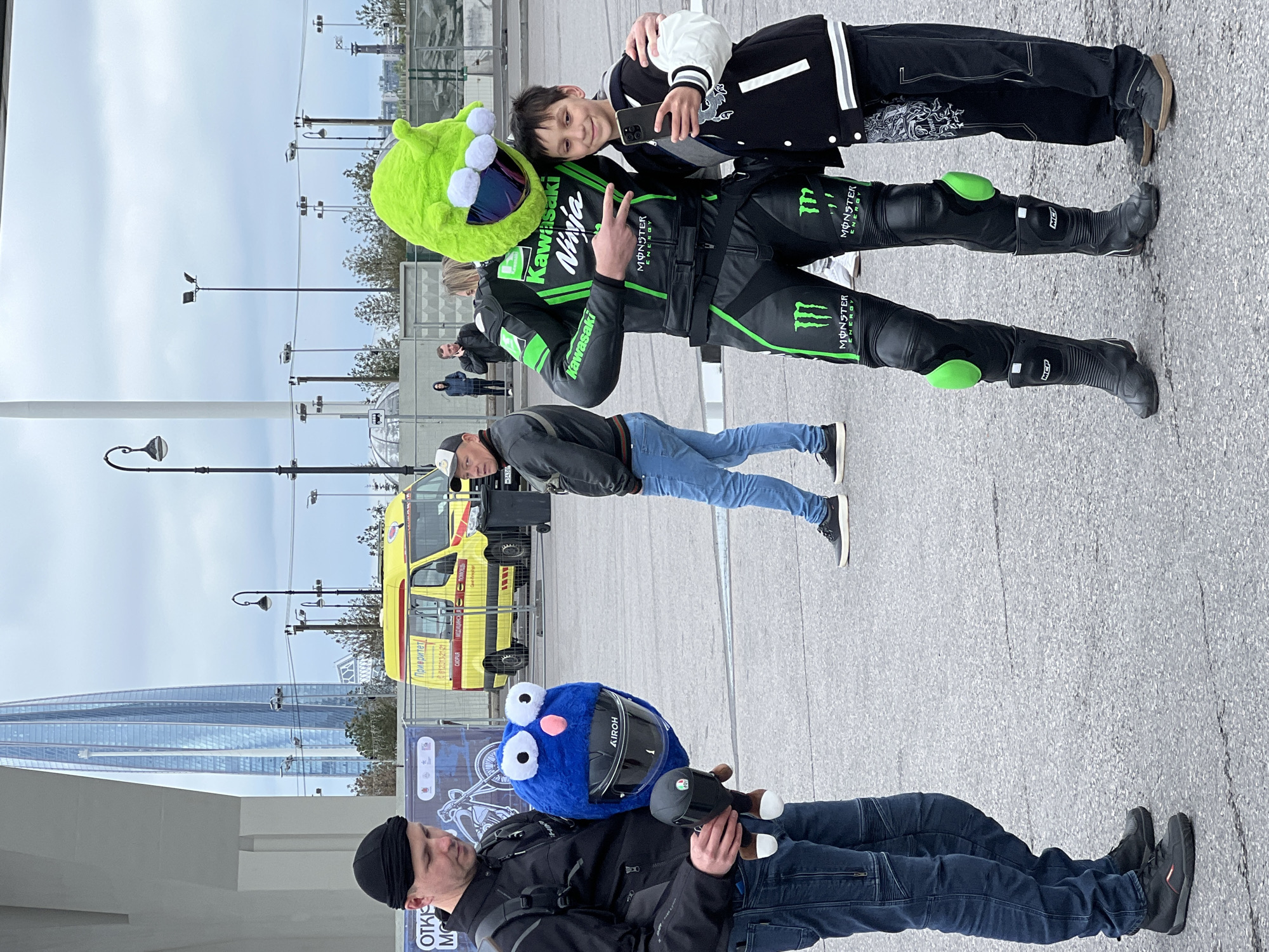 The image shows a street scene likely in Saint Petersburg, Russia, with several people and objects of interest. In the foreground, a man in jeans and a black jacket wearing a black beanie is holding a blue helmet resembling a character. To his right, a motorcyclist wearing a Kawasaki-branded black and green leather suit is making a peace sign, being hugged by a person in a black and white jacket. The motorcyclist is wearing a green helmet resembling a creature. Behind these figures, there is another man wearing blue jeans and a black jacket. In the background, an ambulance with yellow and red striping can be seen, along with streetlights and other people in the distance. A tall, modern skyscraper is visible in the left background. The location is likely a public area with paving, where people have gathered.