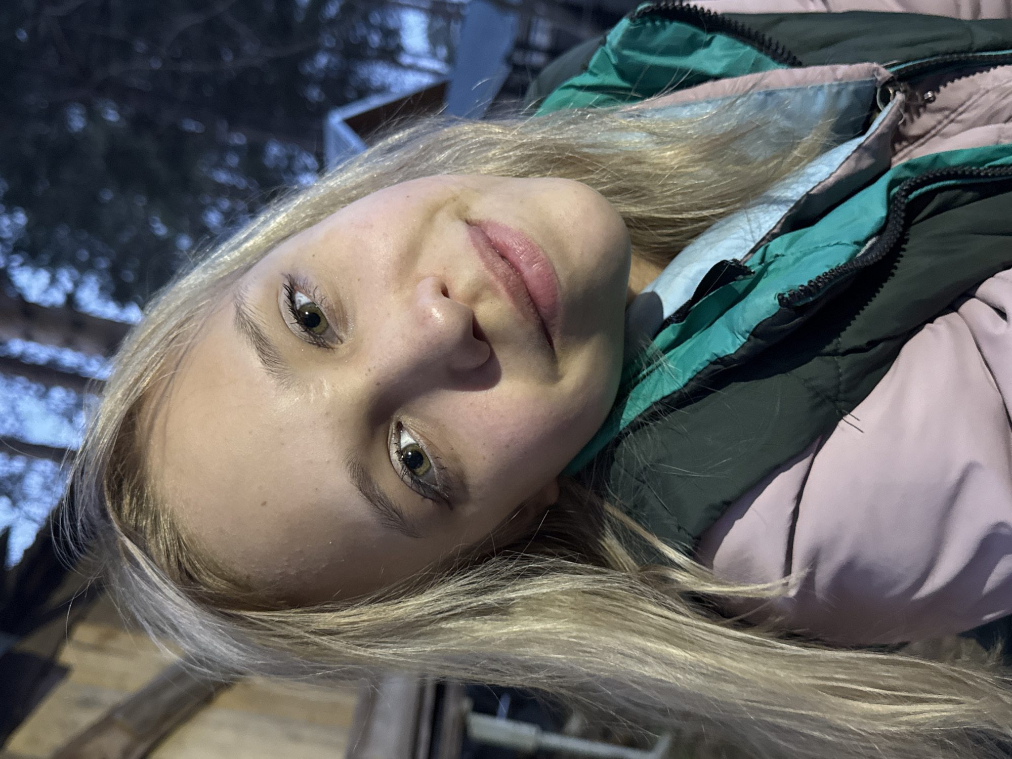 A close-up, portrait-style photograph depicts a young woman outdoors. Her head is tilted to the left. She has fair skin, light blonde hair, and hazel eyes. She is wearing a puffy winter coat in shades of pale pink, green, and black. The background shows hints of trees and a building. The location of Chaikovsky, Russia cannot be verified in the image.