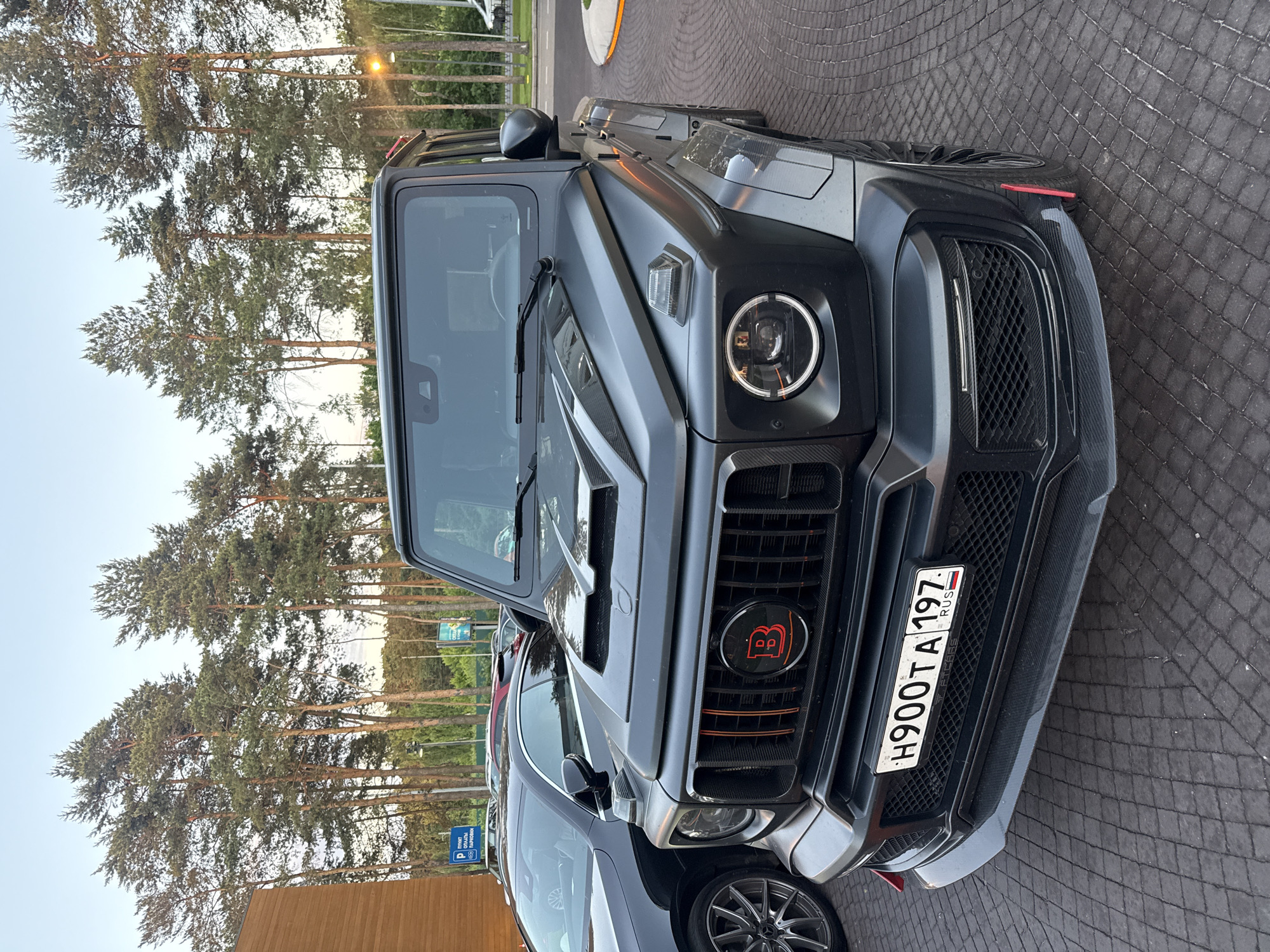An imposing matte black Mercedes-Benz G-Class, modified with a Mansory body kit, is parked on a textured cobblestone surface in front of a wooden building. The vehicle features prominent front bumper modifications, including large air intakes and a carbon fiber splitter, along with a distinctive hood scoop. The front grille has vertical red accents and a red "B" logo, possibly indicating a Brabus modification. The headlights are circular with LED daytime running lights. A Russian license plate, "H900TA 197," is affixed to the front bumper. To the left of the G-Class, a dark-colored sedan with silver alloy wheels is partially visible. Behind the vehicles, a line of tall pine trees forms a backdrop, with a light pole and a blue parking sign visible in the distance. The sky is a clear, pale blue, suggesting it is either early morning or late afternoon. The overall scene appears to be a parking area, possibly near a commercial establishment or a residential complex, in Barvikha, Russia. There are no people visible in the frame.