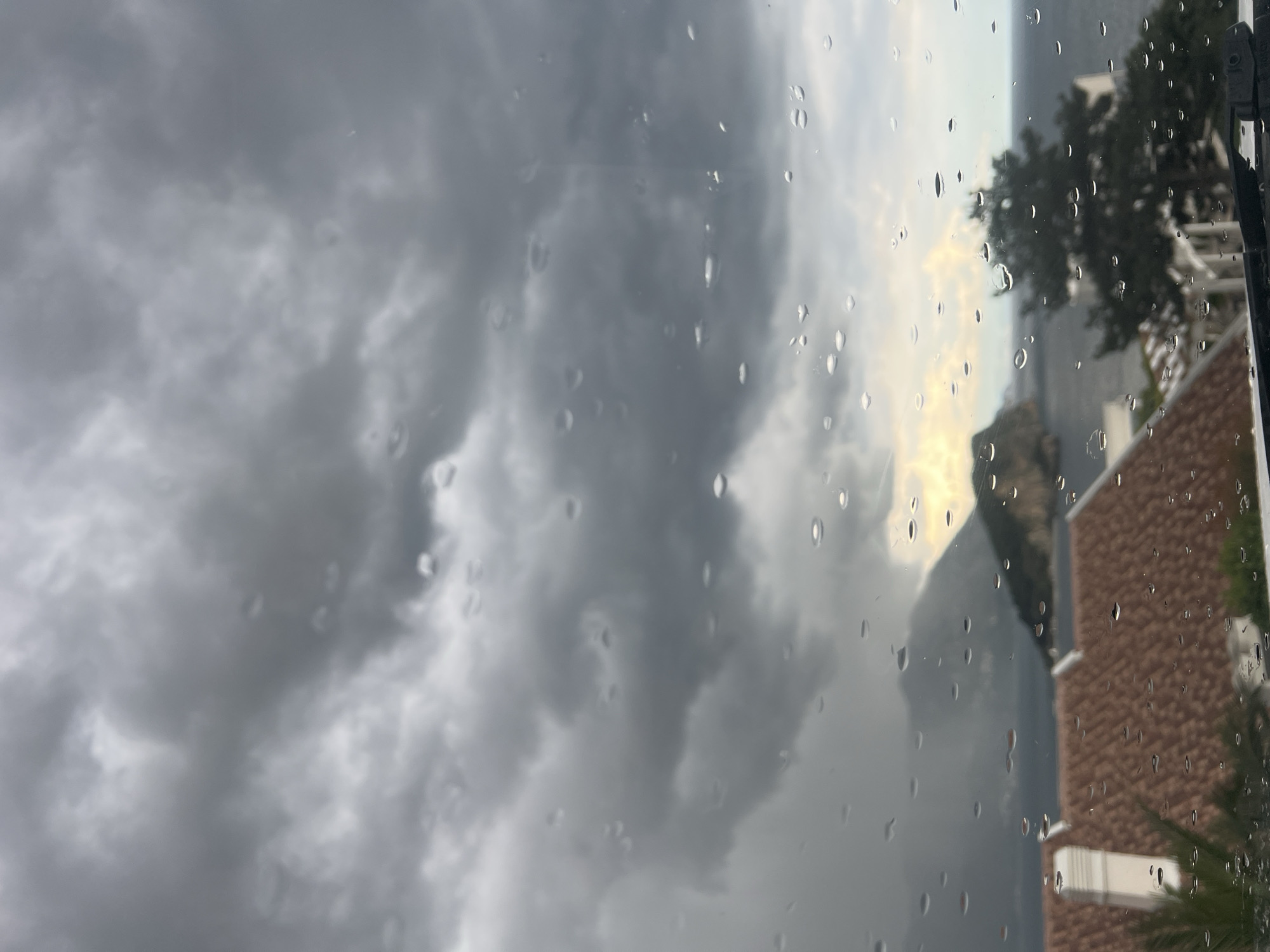 The original image, viewed with the horizon horizontal, captures a dramatic coastal landscape seen through a rain-streaked window. The foreground is dominated by numerous water droplets adhering to the glass, creating a diffuse and textured effect over the distant scene.
The sky is filled with heavy, dark grey, and turbulent clouds, indicating stormy weather or recent rainfall. Towards the center-right, a narrow strip of brighter, yellowish-white sky is visible, suggesting either a temporary break in the clouds or the light from a setting or rising sun attempting to pierce through.
In the midground, a large, dark mountainous landmass rises prominently. Below it, a dark, somewhat reflective horizontal expanse is visible, strongly suggesting the sea. On the right side of the frame, parts of human structures are present, including a building with a reddish-brown, terracotta-tiled roof and white architectural details, typical of coastal Mediterranean regions. Dark green trees and foliage are scattered across the visible land, with a distinct palm-like frond visible at the bottom right.
The image conveys the atmosphere of a rainy or recently rained-upon day, viewed from a sheltered indoor position. The dominant weather is overcast and cloudy. The architecture and mountainous coastline are consistent with the provided context of Budva, Montenegro. No text is visible in the media.
