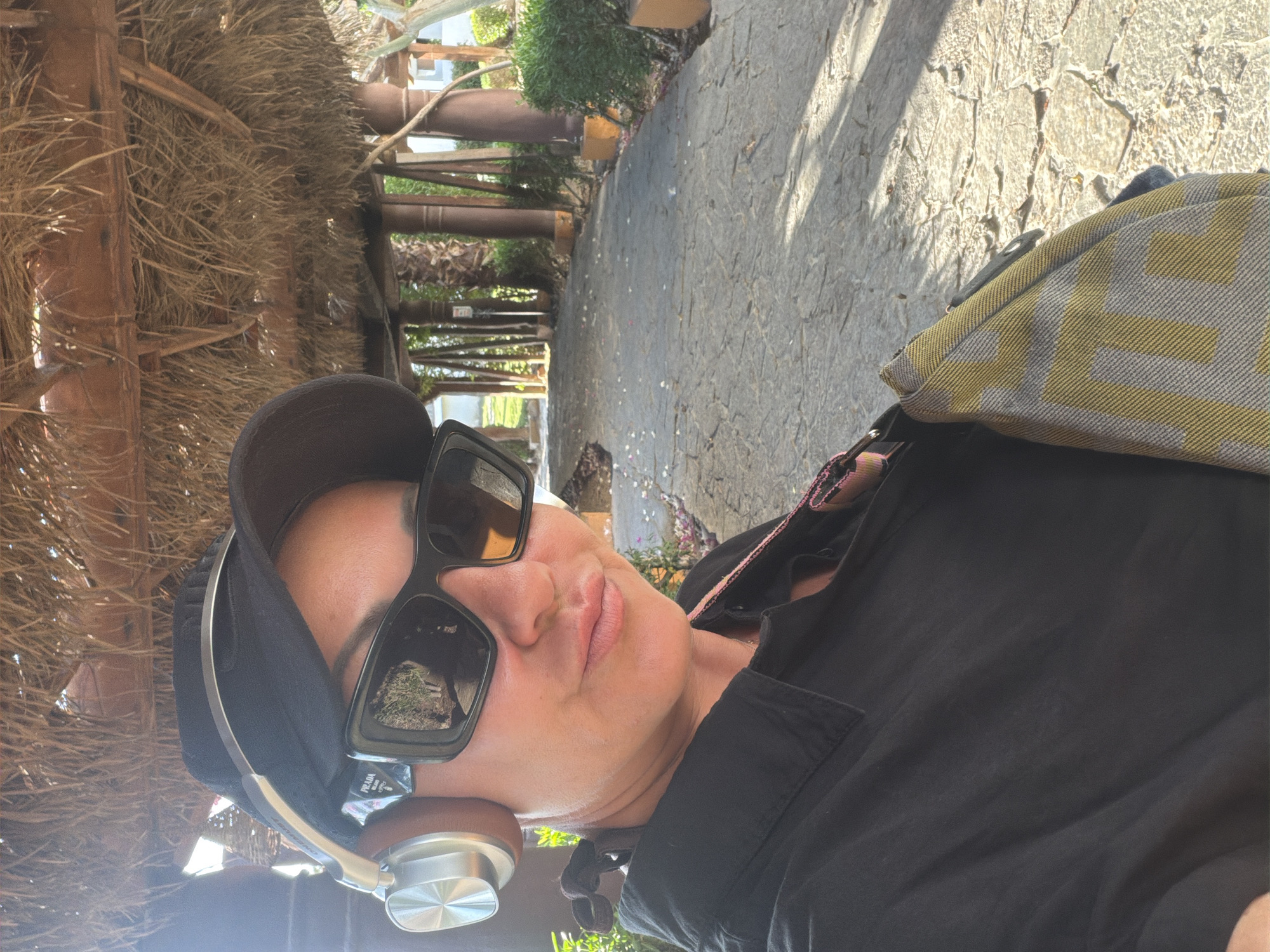 A woman wearing a black baseball cap, black sunglasses, and a black collared shirt takes a selfie under a thatched roof structure. She has silver and brown headphones on and a patterned bag strap visible. The background shows a paved walkway lined with palm trees and bushes, suggesting an outdoor setting in Sharm El Sheikh, Egypt, possibly a resort area given the architectural style of the roof and surrounding vegetation. The sun casts shadows on the ground.